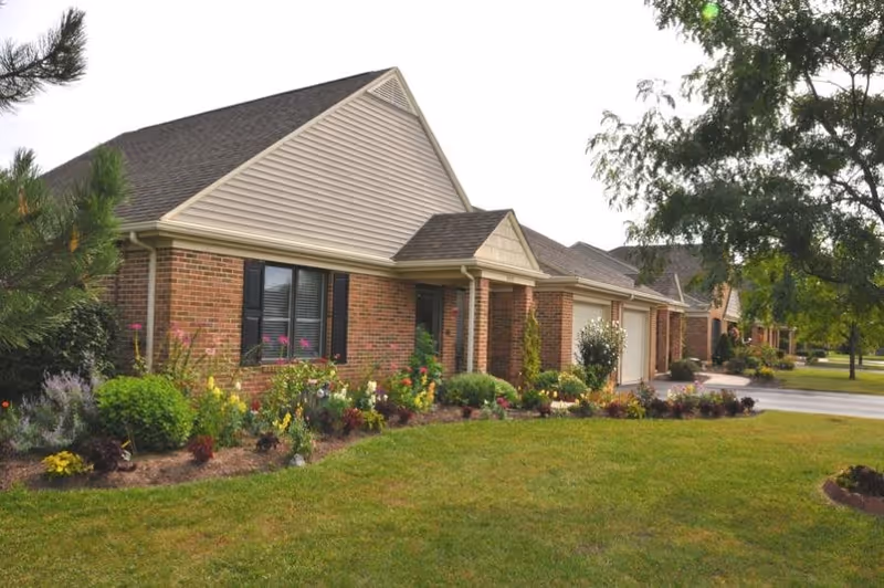 Exterior view of a single-story brick senior living community building with a well-maintained garden featuring various colorful flowers and shrubs in front, a driveway, and trees in the background.
