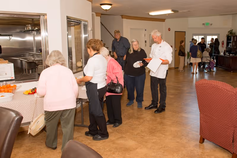 A group of elderly people and staff members standing in line at a serving counter inside a senior living facility. The room has a tiled floor, beige walls, and a few chairs and tables. Some people are interacting with a man holding a clipboard, and others are waiting in line near a kitchen pass-through window.