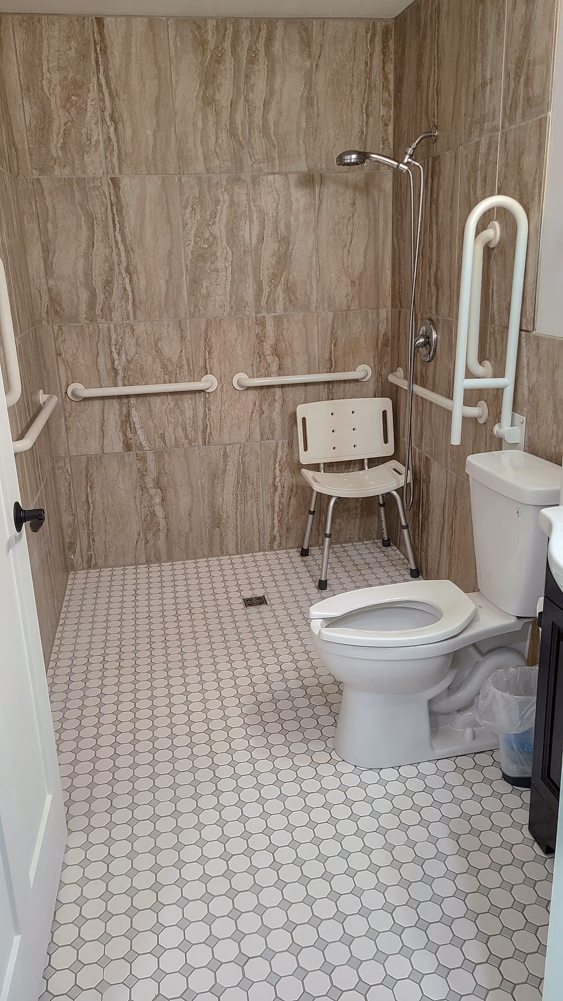 Accessible bathroom with a toilet, shower chair, wall-mounted showerhead, and multiple grab bars over tiled walls and hexagonal tile floor.