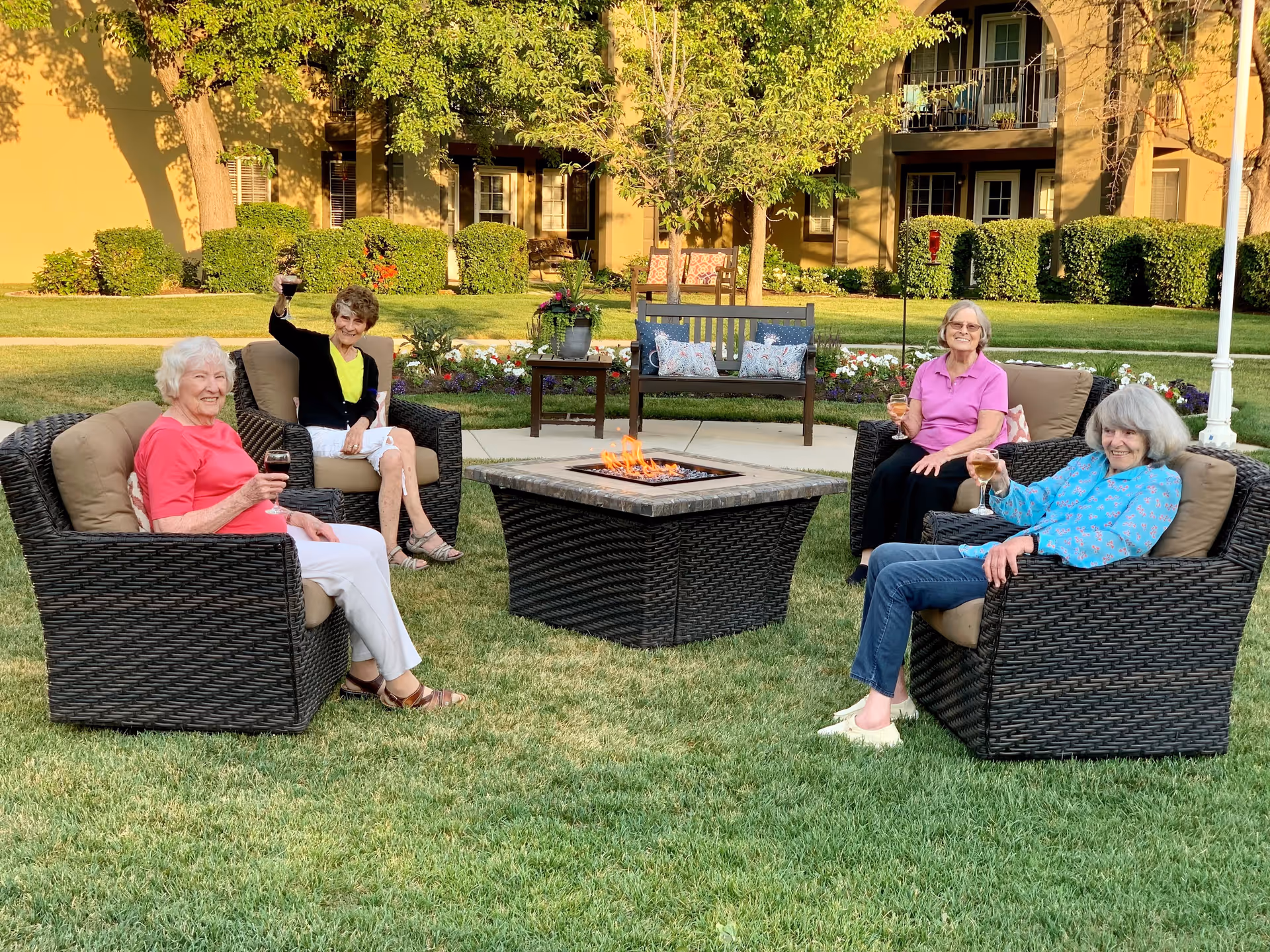 Four elderly women sitting outdoors in wicker chairs arranged around a square fire pit on a grassy lawn, each holding a glass and smiling. Behind them are trees, flower beds, and a building with balconies and windows.