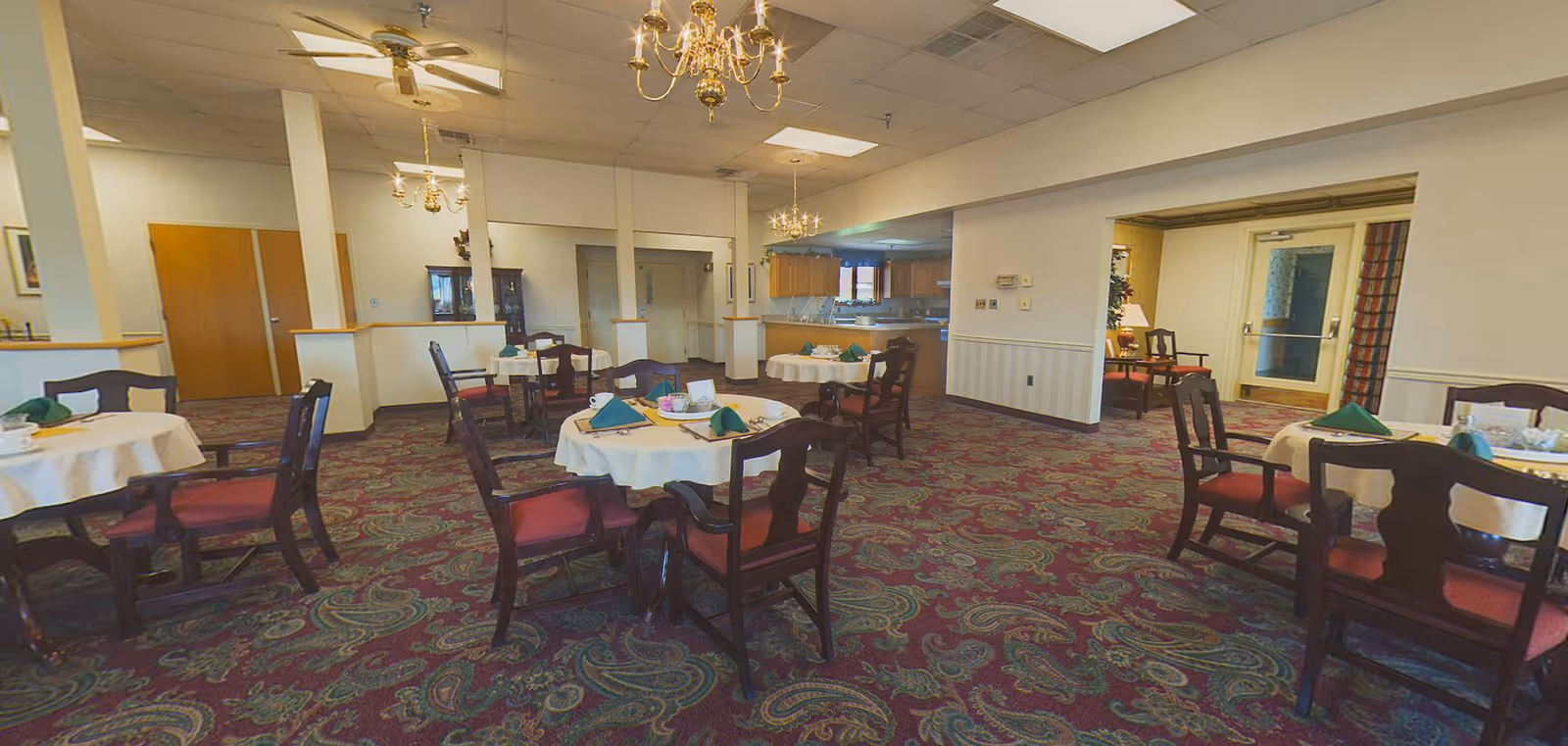 A dining room with round tables covered with white tablecloths and set with green napkins, cups, and utensils. The room has patterned carpet, wooden chairs with red cushions, chandeliers, ceiling fans, and a kitchen area in the background.