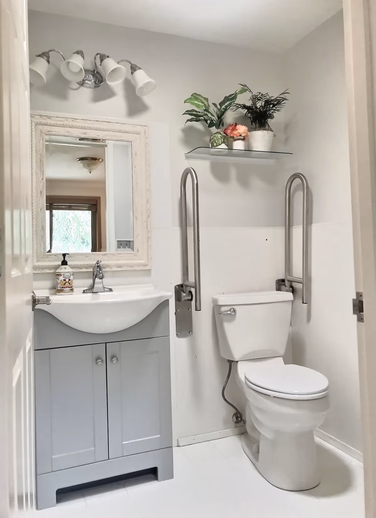 A clean and bright bathroom with a white toilet equipped with two stainless steel grab bars on either side. Next to the toilet is a gray vanity with a white sink and a soap dispenser. Above the sink is a white-framed mirror and a light fixture with four downward-facing lamps. A glass shelf above the toilet holds three potted plants.