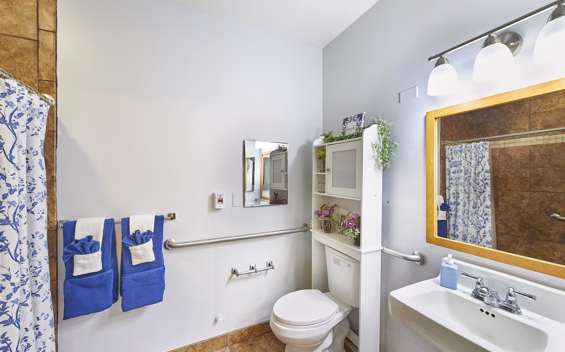 Well-lit bathroom featuring a toilet and sink, a shower with a blue floral curtain, blue folded towels, shelving above the toilet, and wall-mounted grab bars.