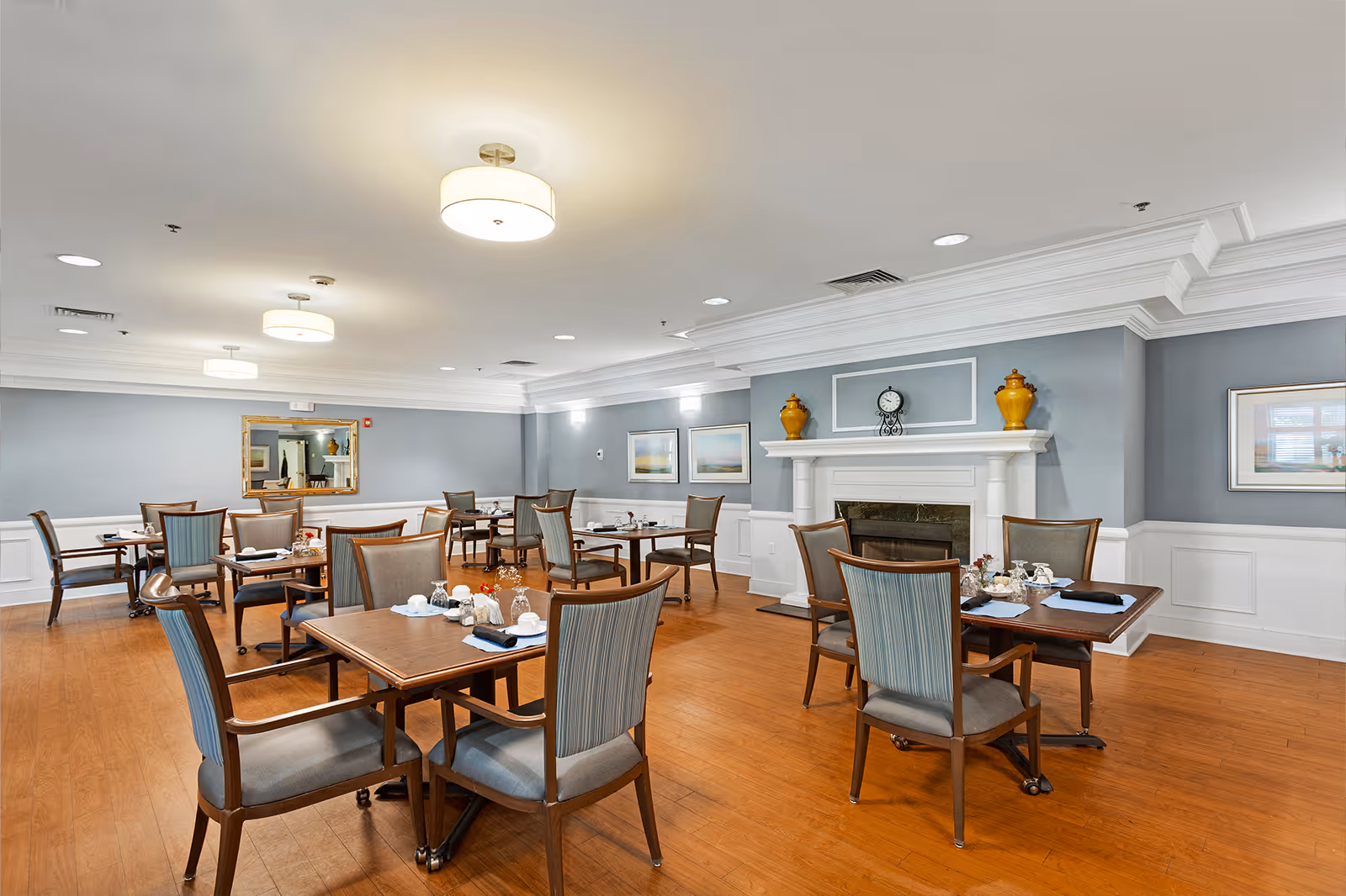 A spacious dining room with several wooden tables and cushioned chairs arranged neatly. The room features light blue walls with white wainscoting, a wooden floor, and a white fireplace adorned with a clock and decorative vases. Ceiling lights provide warm illumination, and framed artwork hangs on the walls.