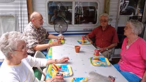 Four elderly people sitting around a table outdoors, enjoying a meal together with colorful plates and soda cans on the table. They are engaged in conversation and appear to be in a relaxed social setting.