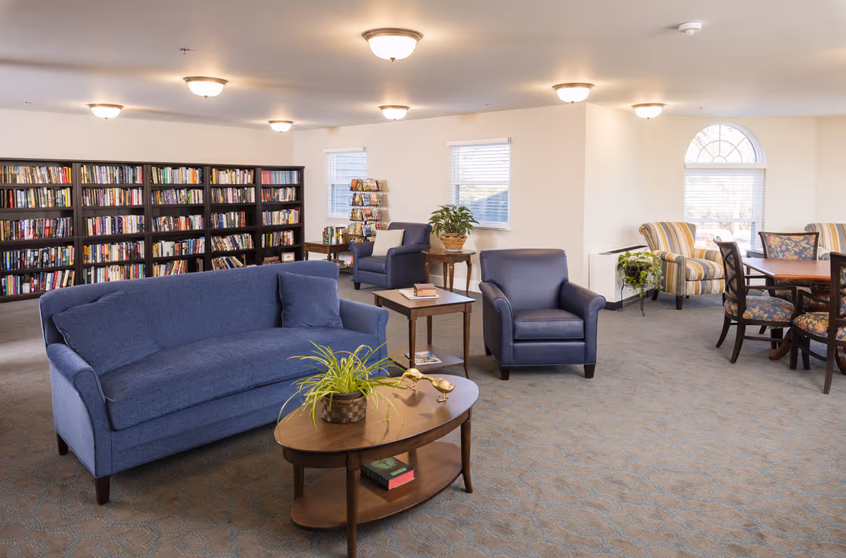 A spacious and well-lit common area with blue upholstered sofa and armchair, wooden coffee and side tables with plants and books, a large bookshelf filled with books along the back wall, additional seating with striped armchairs near windows, and a round table with floral cushioned chairs.