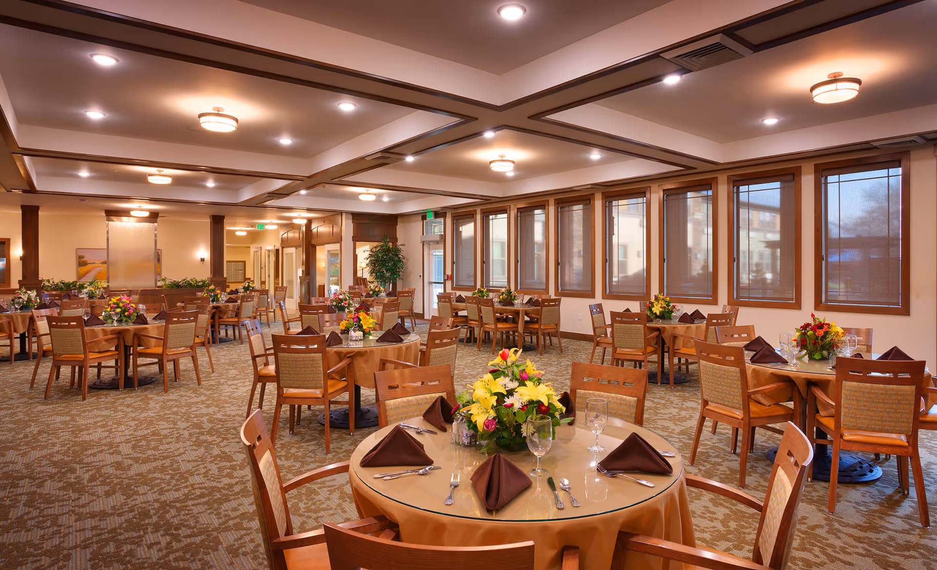 A spacious senior living dining room with multiple round tables covered with beige tablecloths, each set with brown folded napkins, silverware, glassware, and floral centerpieces. The room has large windows with blinds, warm lighting from ceiling fixtures, and a carpeted floor.