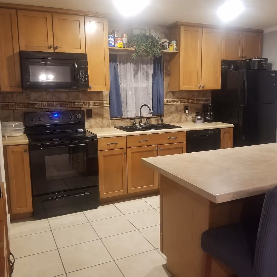 A kitchen with wood cabinets, black appliances, a sink under a small curtained window and a center island.