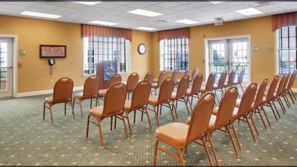 A conference or meeting room with rows of brown chairs arranged facing a podium. The room has yellow walls, large windows with blinds, and double glass doors leading outside. The floor is carpeted in green with a subtle pattern.