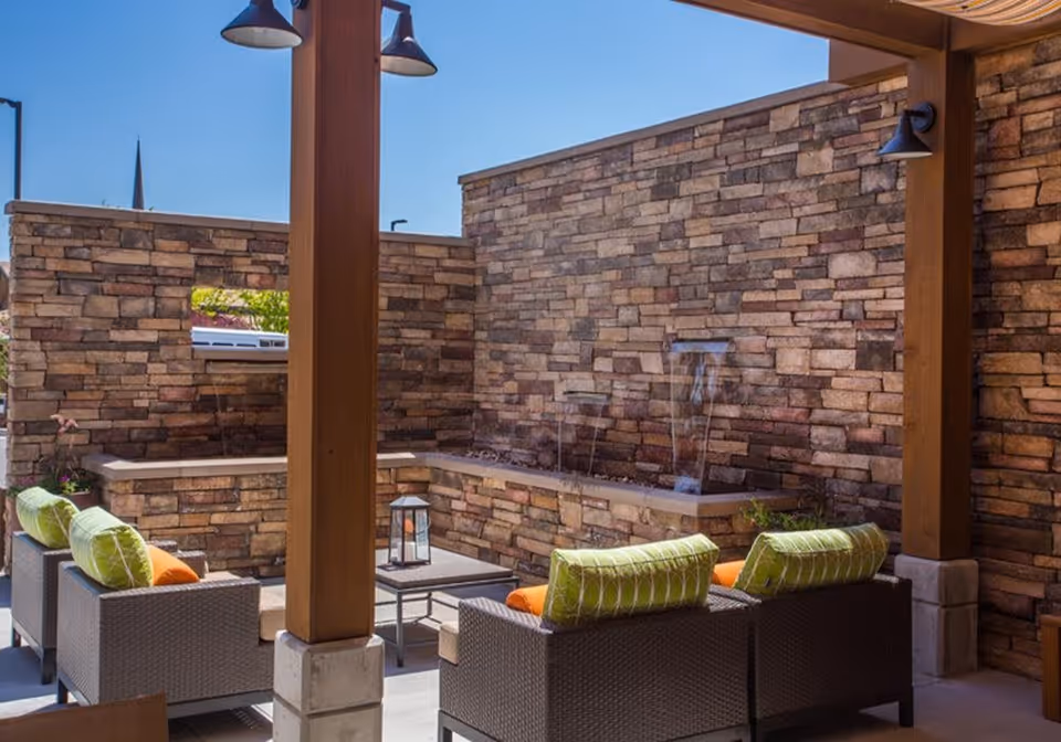 Outdoor patio area with wicker seating featuring green and orange cushions, a small table with a lantern, and a stone wall with a water feature under a wooden pergola with overhead lights.