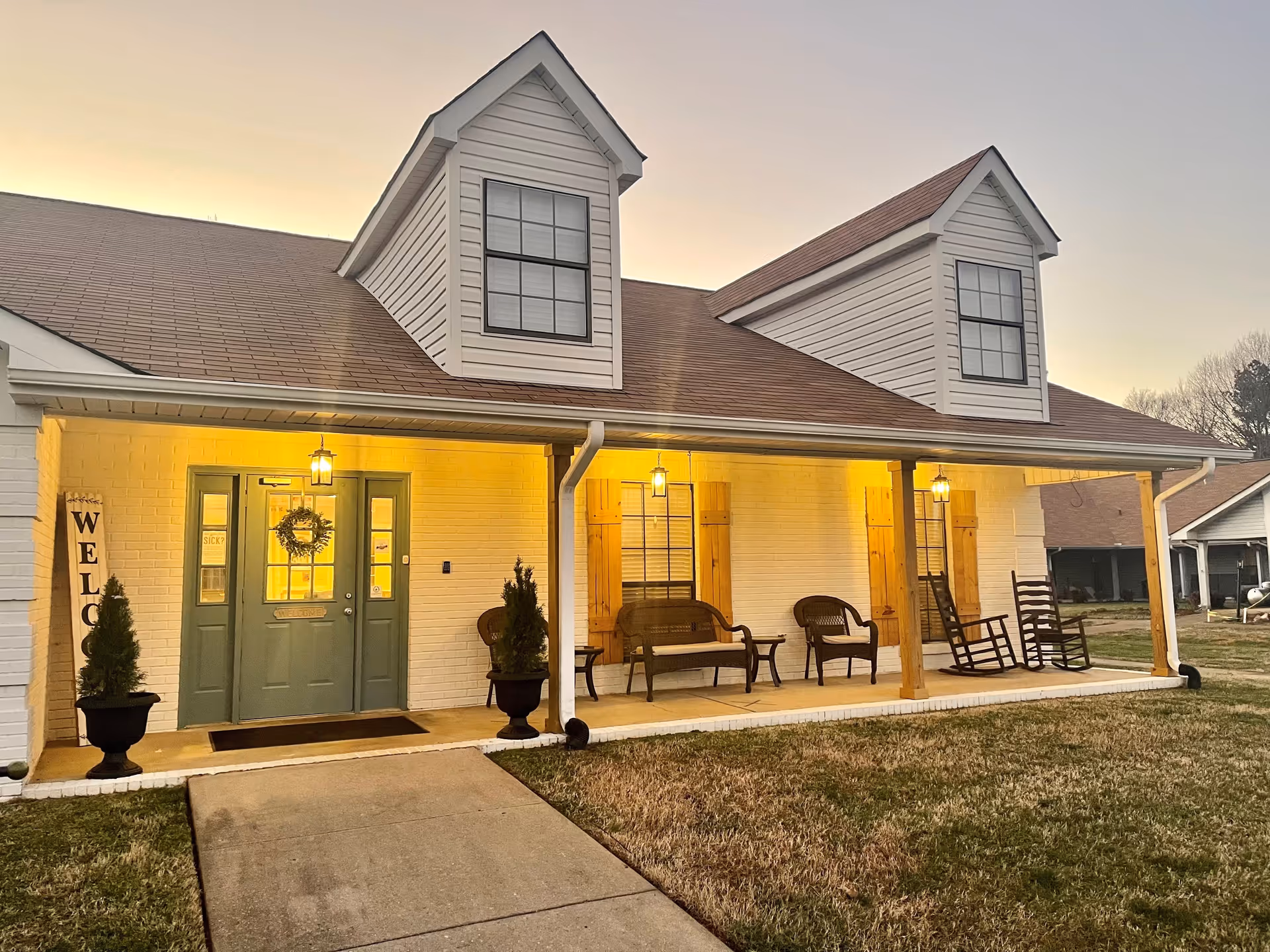 Exterior view of a single-story building with a porch at dusk. The building has white brick walls, a brown shingled roof with two dormer windows, and a green double door entrance with a wreath. The porch is lit with warm yellow lights and features wooden rocking chairs and benches. Two potted plants flank the entrance, and a vertical 'WELCOME' sign is displayed on the left side of the door.