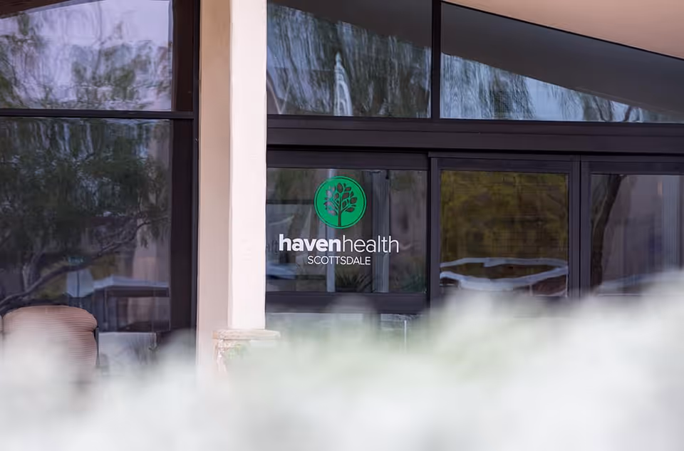 Glass entrance door of a building with the logo and text 'haven health SCOTTSDALE' displayed on the glass. There are reflections of trees and outdoor surroundings visible on the glass panels.