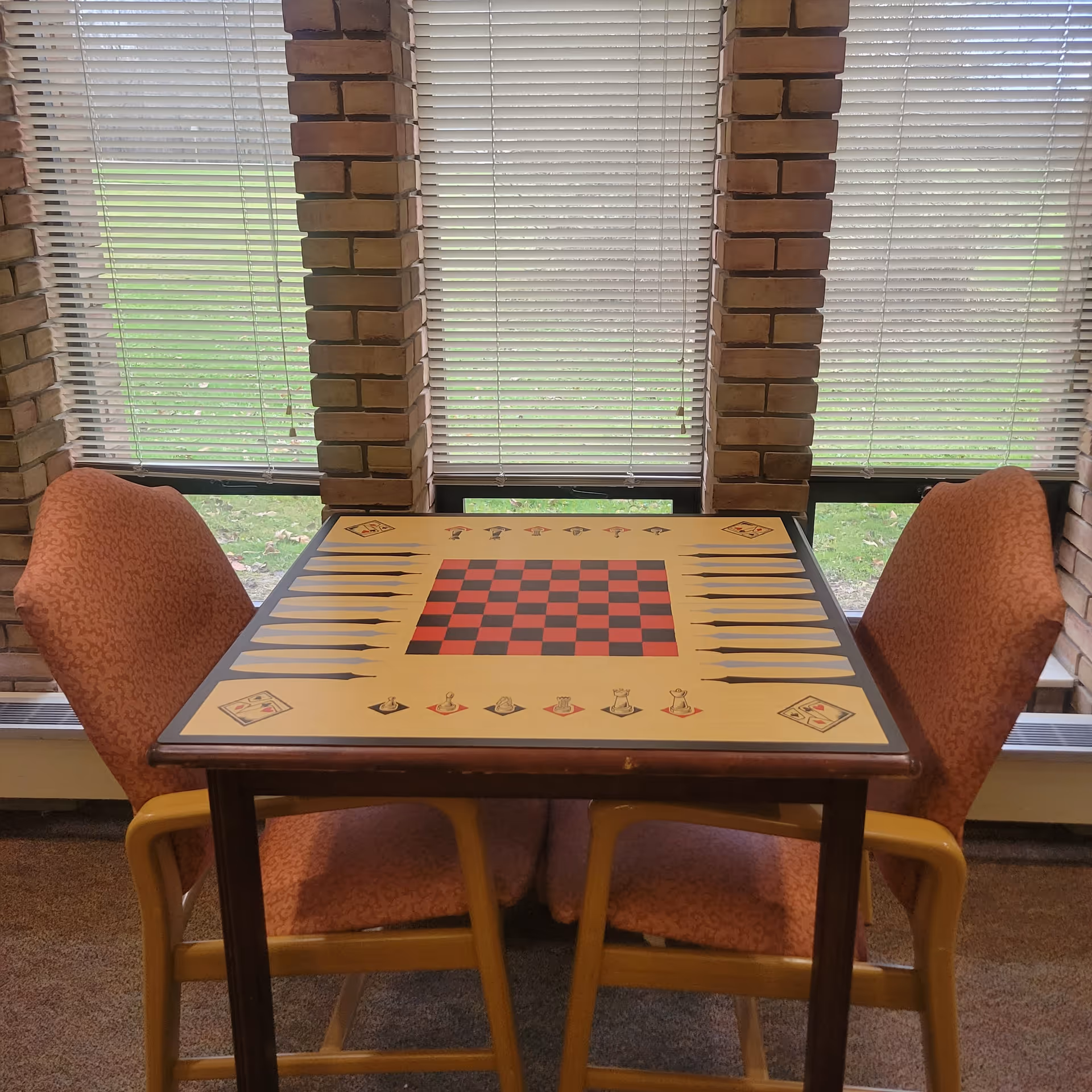 A small square game table with a checkerboard and backgammon design on the surface, flanked by two orange cushioned chairs with wooden armrests. The table is positioned in front of three windows with closed white blinds, framed by brick columns. Outside the windows, green grass is visible.