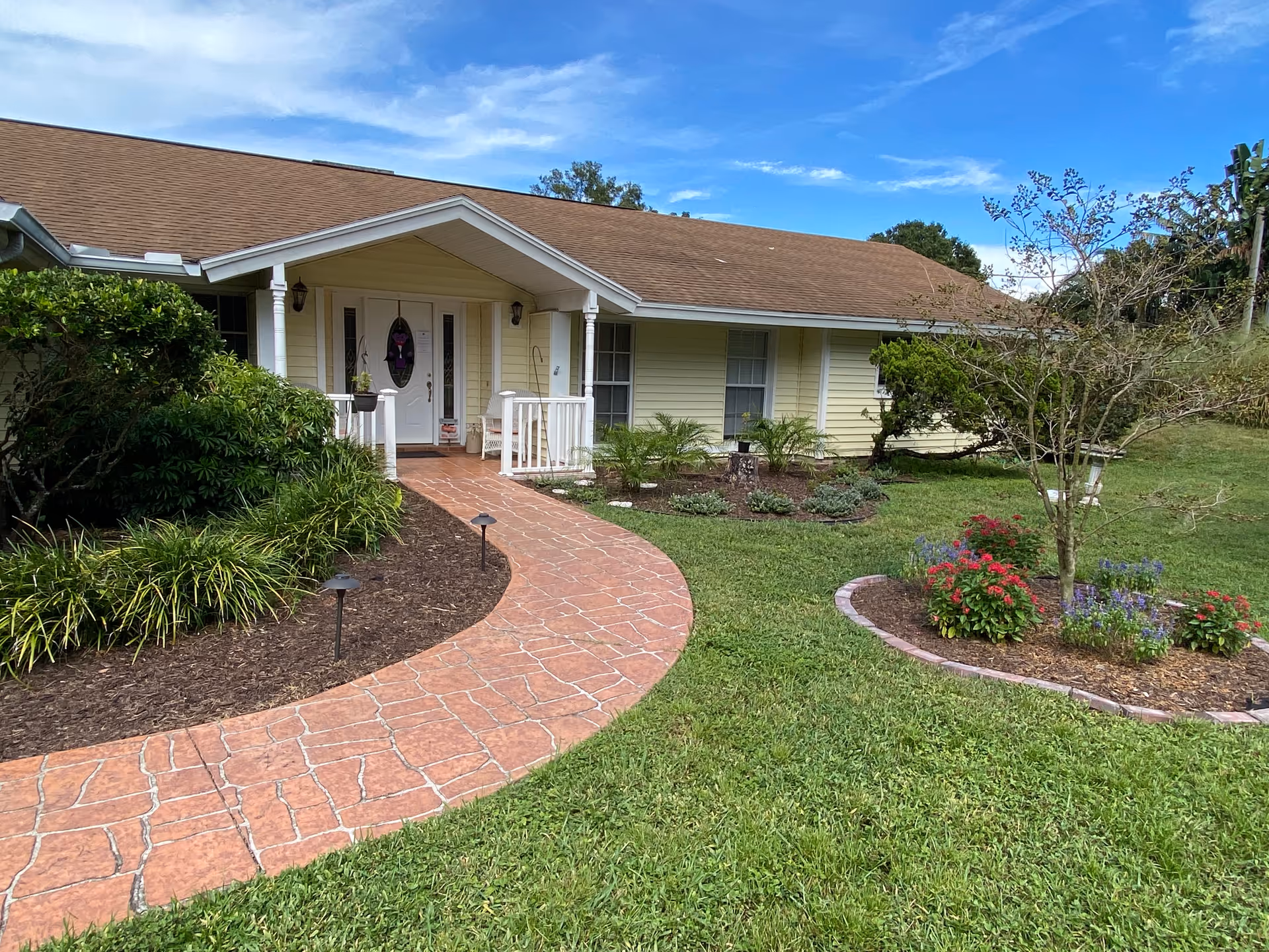 Front exterior view of a single-story yellow building with a brown shingled roof, a white front door with glass panels, a curved brick walkway leading to the entrance, and landscaped garden beds with green shrubs, small trees, and colorful flowers under a partly cloudy blue sky.