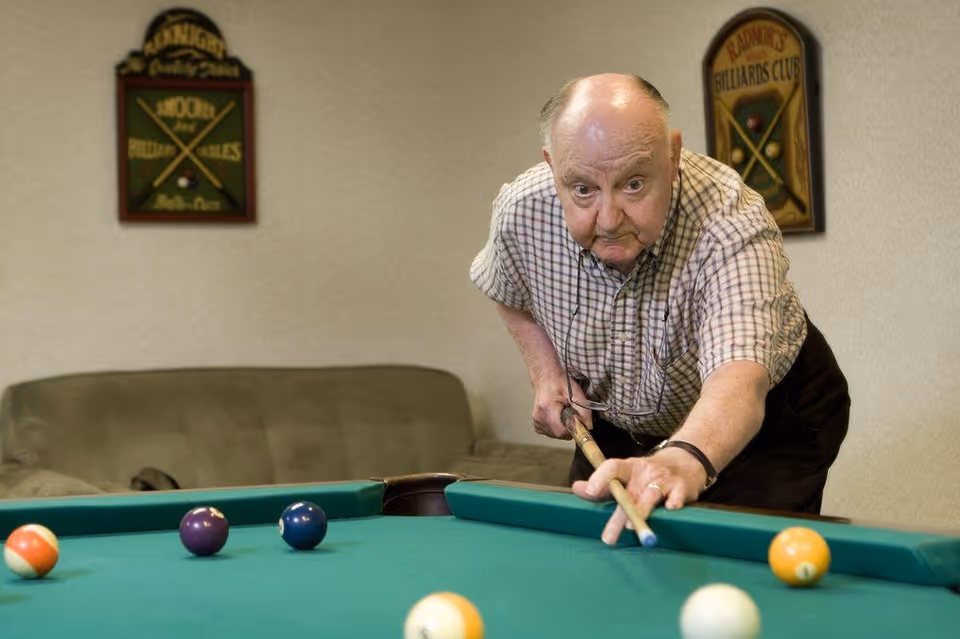 An elderly man playing billiards indoors, leaning over a pool table and aiming with a cue stick. Behind him is a green couch and two wall plaques related to billiards.