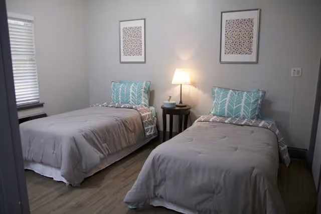 Two twin beds in a simple bedroom with a shared bedside table and lamp.