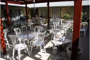 Outdoor patio area with multiple white metal tables and chairs arranged under a wooden pergola. The space is surrounded by windows and walls of a building, with some potted plants and decorative elements visible.