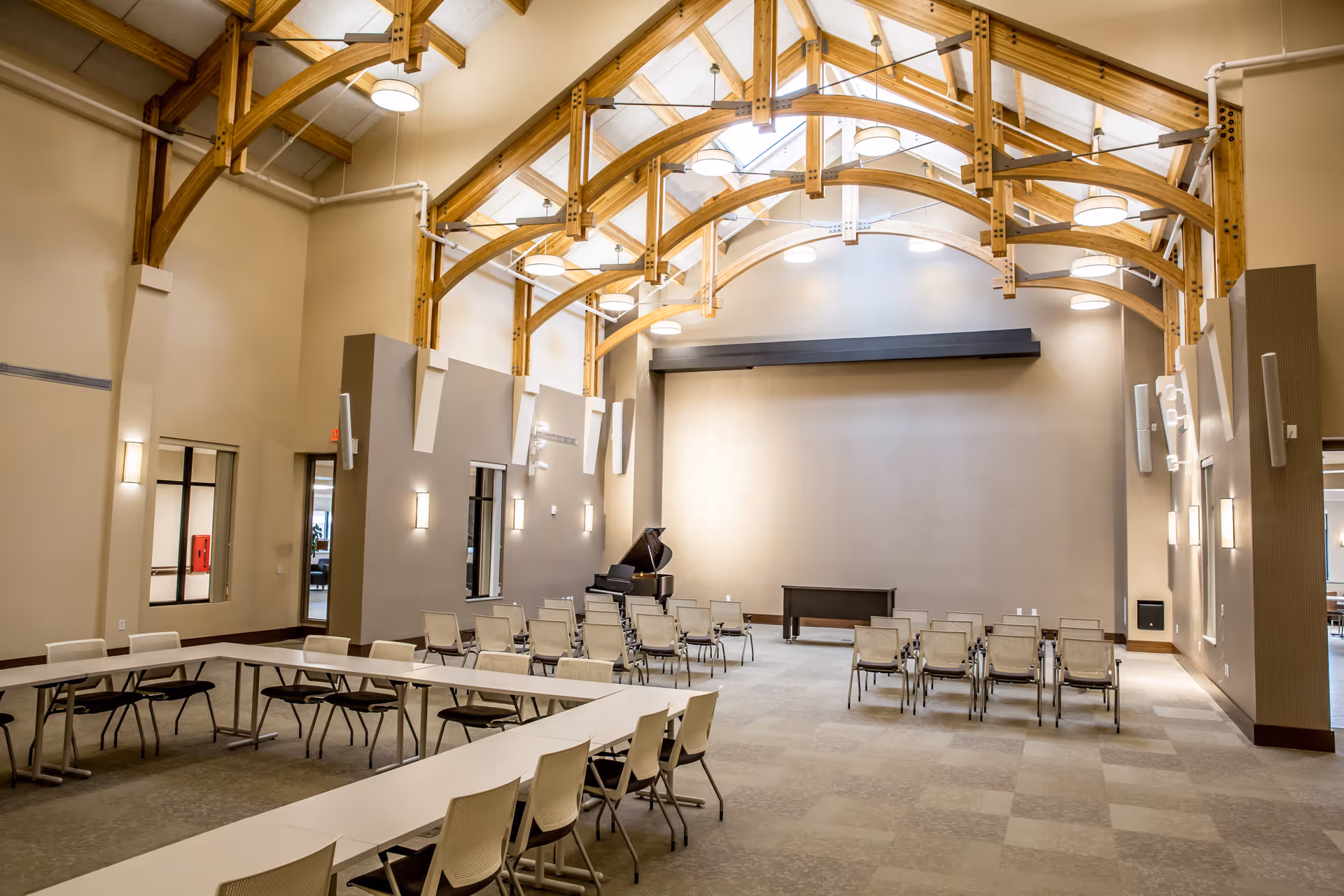 A spacious room with high vaulted ceilings featuring wooden beams and multiple round ceiling lights. The room has beige walls and carpeted floors. There are several rows of chairs arranged facing a small stage area with a black grand piano and a black table. In the foreground, there are white tables arranged in a U-shape with white chairs around them. The room appears to be a multipurpose gathering or event space.