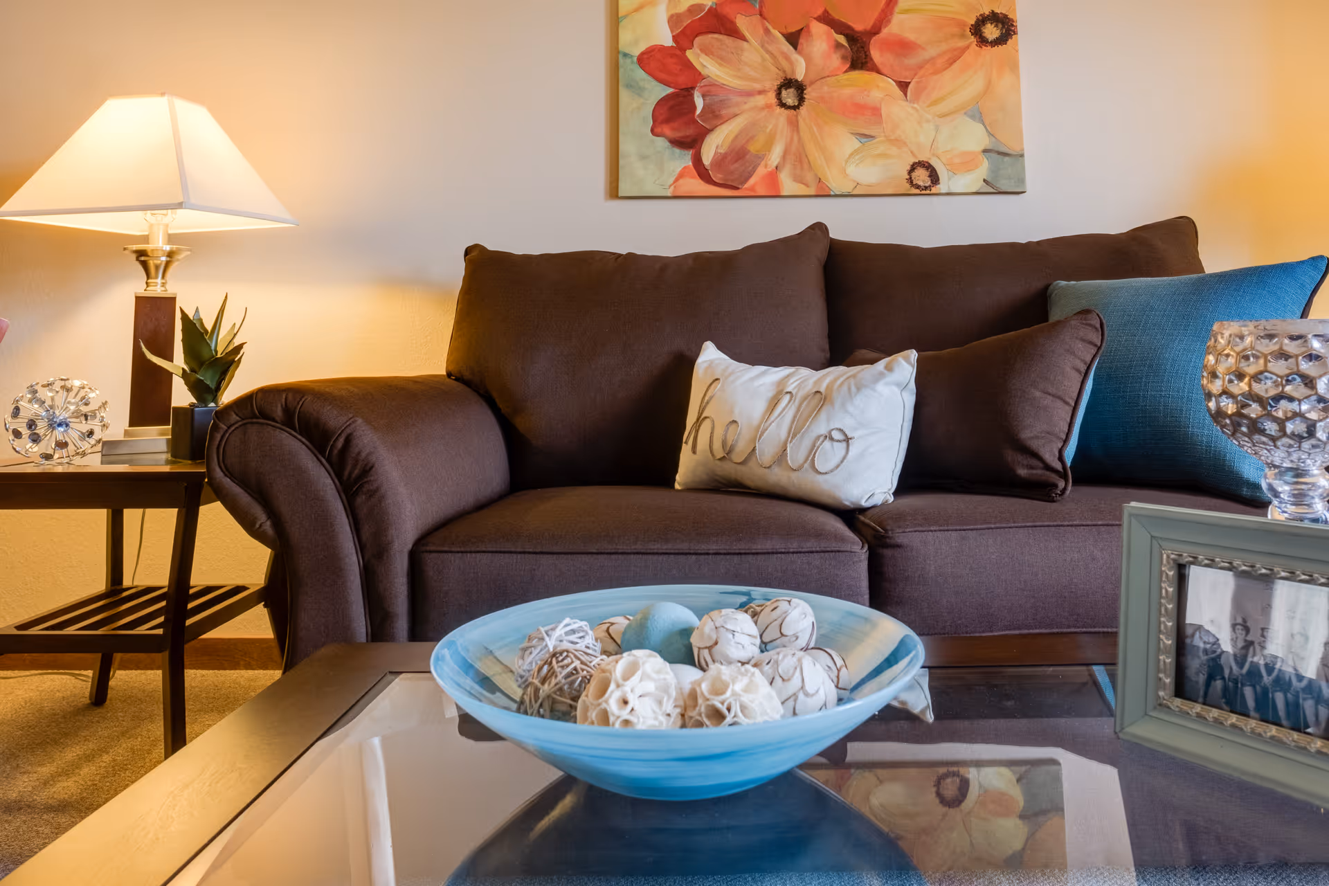 Cozy living room with a brown sofa adorned with three pillows, one of which has the word 'hello' embroidered on it. A glass coffee table in front holds a decorative blue bowl filled with assorted decorative balls. To the left, there is a side table with a lamp, a small plant, and a decorative object. A colorful floral painting hangs on the wall above the sofa.