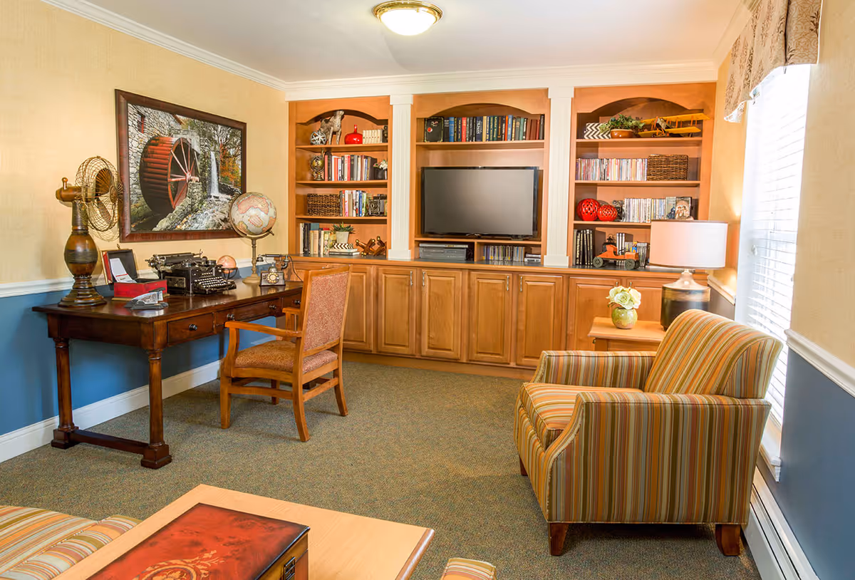 Cozy common room with built-in wooden bookshelves and TV, a desk with a globe and vintage typewriter, and striped armchairs by a window.