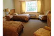 A nursing home bedroom with two single beds covered in brown bedspreads, a wooden nightstand with a lamp between the beds, a window with yellow curtains, a cushioned armchair, a wooden dresser, and a carpeted floor.