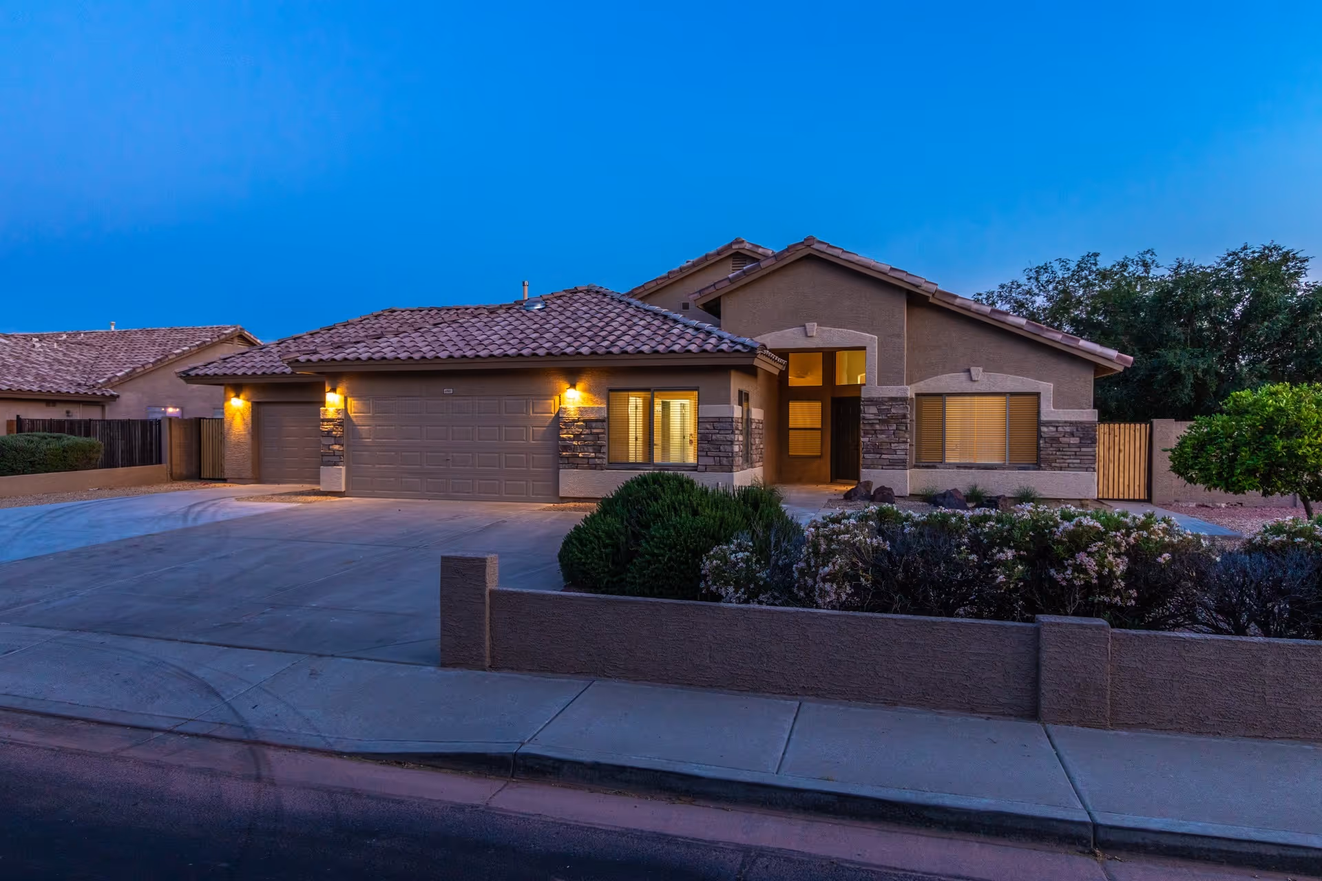 Exterior view of a single-story house with a tiled roof and a three-car garage at dusk. The house has stone accents on the facade, warm exterior lights near the garage and entrance, and a landscaped front yard with bushes and small trees. The sky is clear and blue.