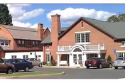 Exterior view of a large brick building with multiple windows and a green roof, featuring a white entrance with columns and a small balcony above. Several cars are parked in front of the building under a partly cloudy sky.