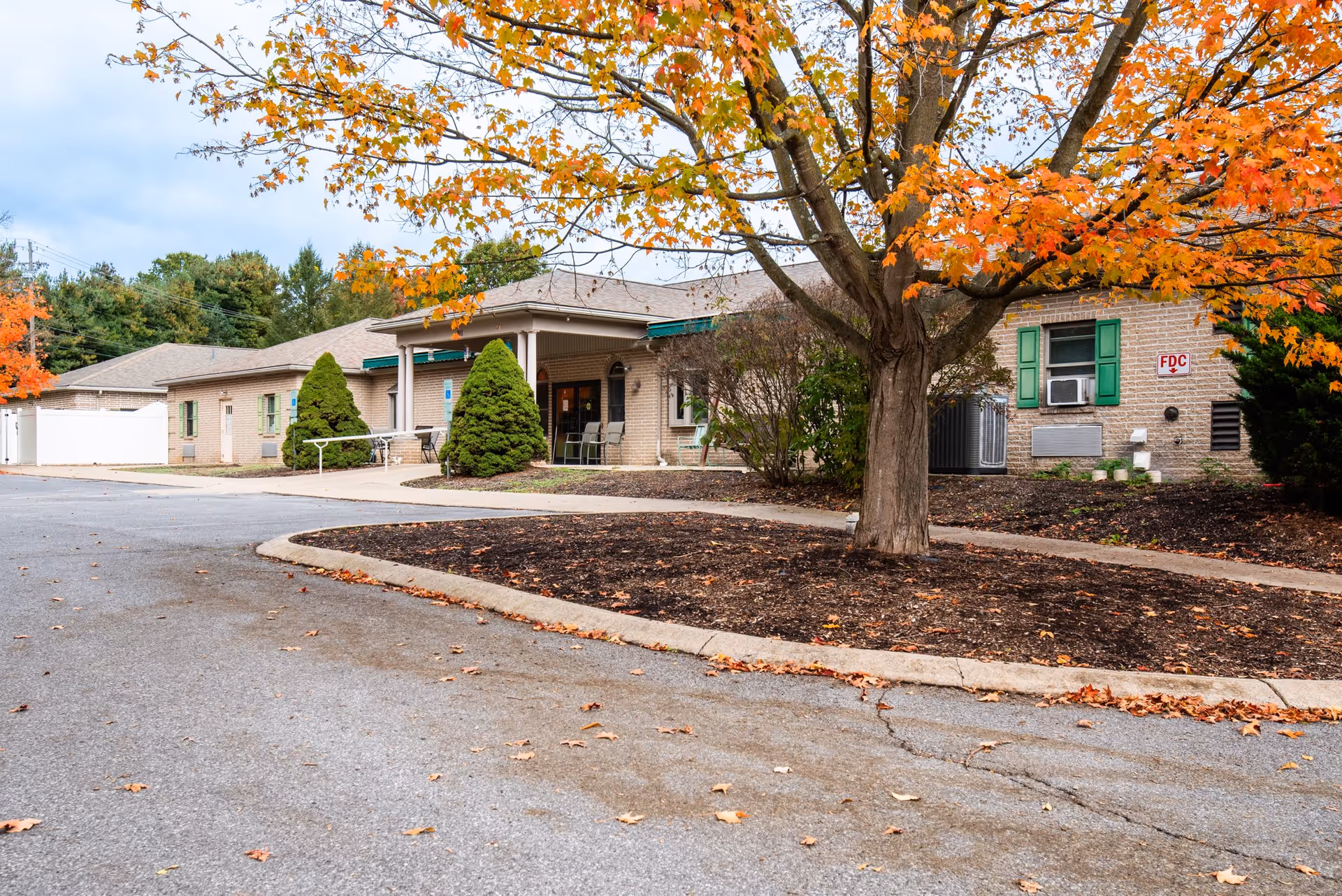 Front entrance of a single-story brick senior living building with an autumn tree, shrubs, and a curved driveway.