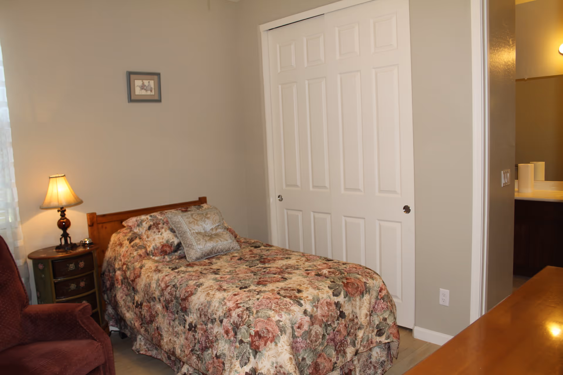 A small bedroom with a single bed covered in a floral bedspread and a decorative pillow. Next to the bed is a round wooden nightstand with a lamp and a small bell. There is a maroon upholstered chair in the corner near a window with sheer white curtains. A white sliding closet door is visible on the wall opposite the bed, and a doorway leads to a bathroom with a sink and mirror.
