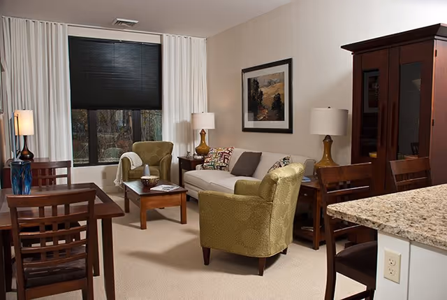 Furnished living room featuring a sofa, two green armchairs, coffee table, dining table and wooden cabinet under a large window.