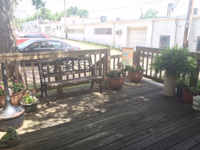 Wooden outdoor deck with a bench, potted plants, and railing overlooking a street with parked cars.