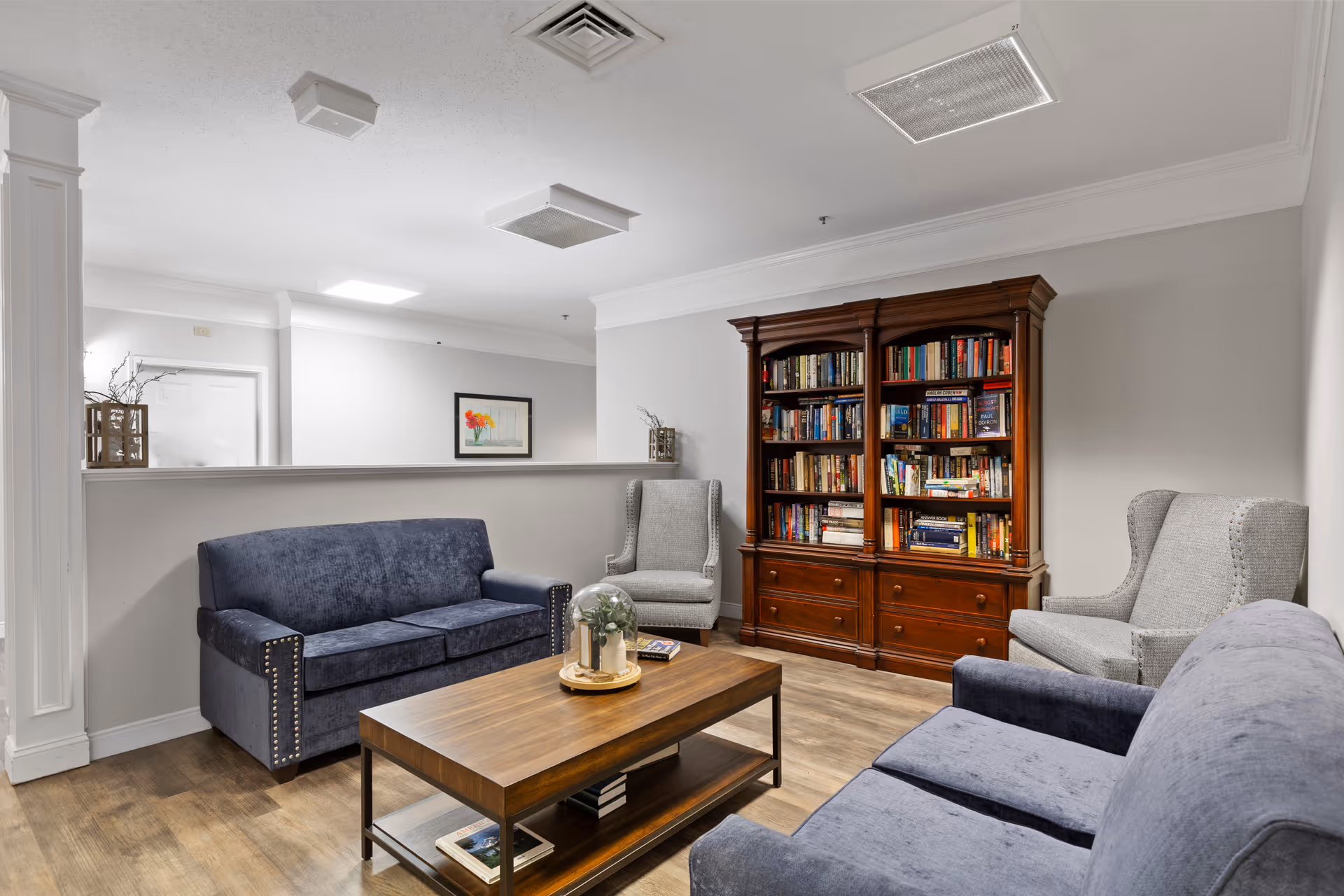 A cozy living room area with two blue upholstered sofas, two gray armchairs, a wooden coffee table with a decorative centerpiece, and a large wooden bookshelf filled with books against a light gray wall.