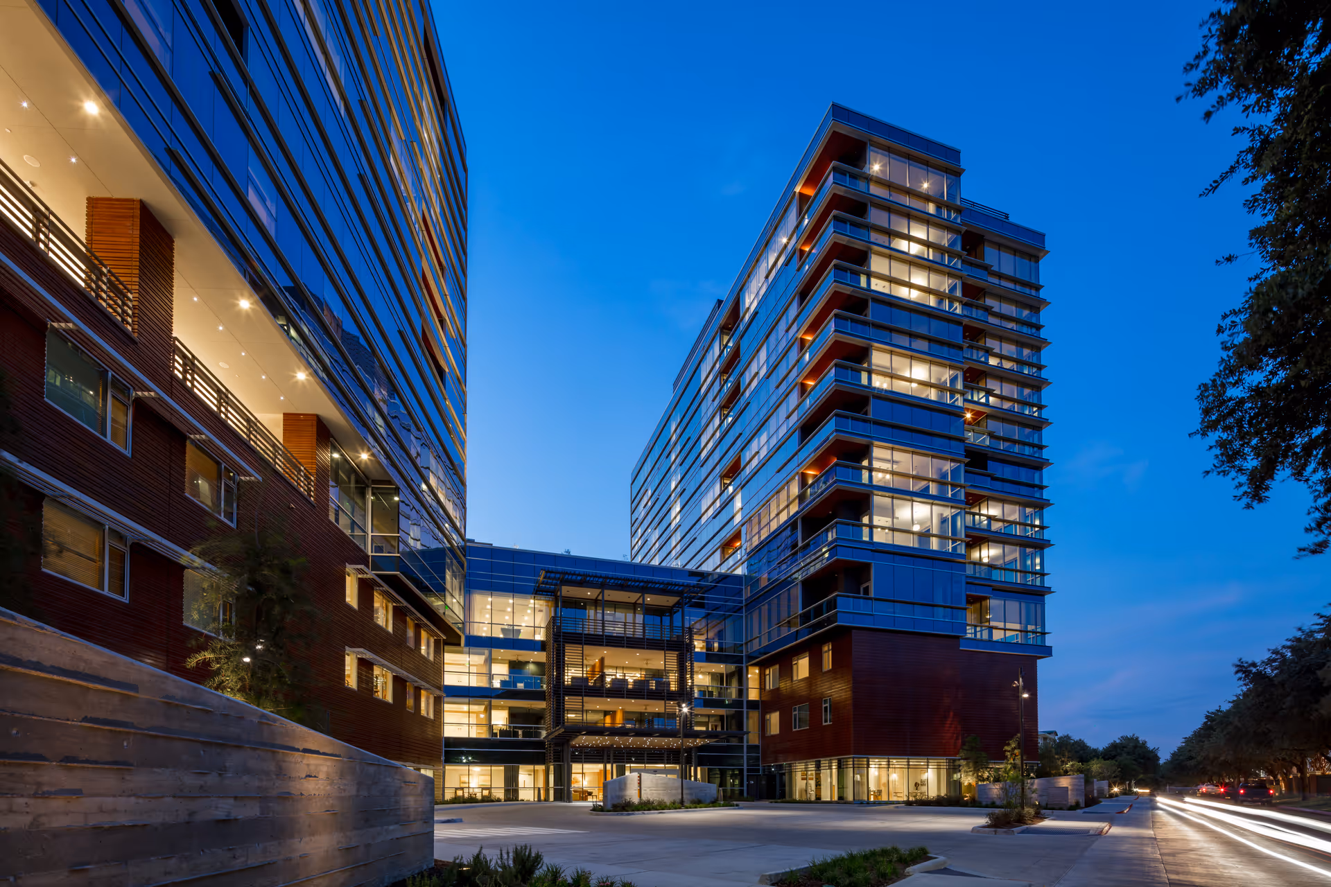 Modern multi-story building with glass windows and illuminated interiors at dusk, featuring a spacious entrance area and surrounded by trees and a clear evening sky.