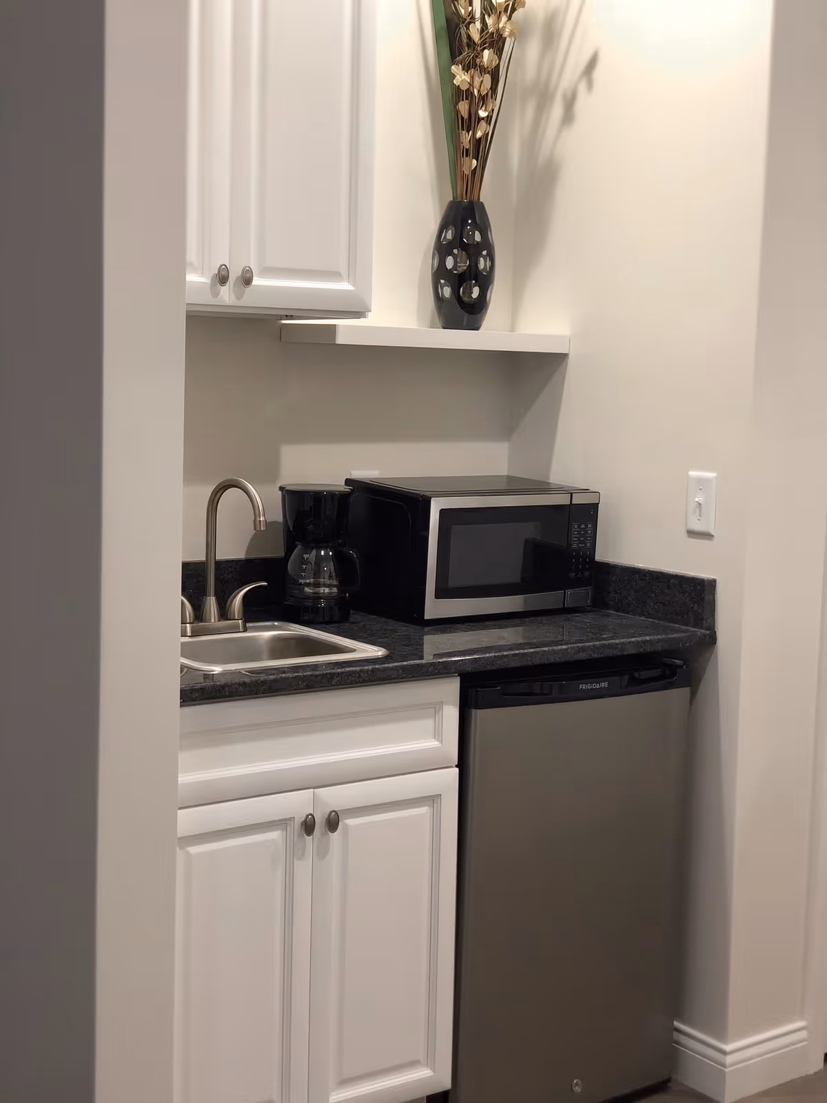 A small kitchenette area with white cabinets, a black countertop, a stainless steel sink with a faucet, a black coffee maker, a black microwave, and a stainless steel mini refrigerator. A decorative black vase with dried flowers is placed on a white shelf above the microwave.