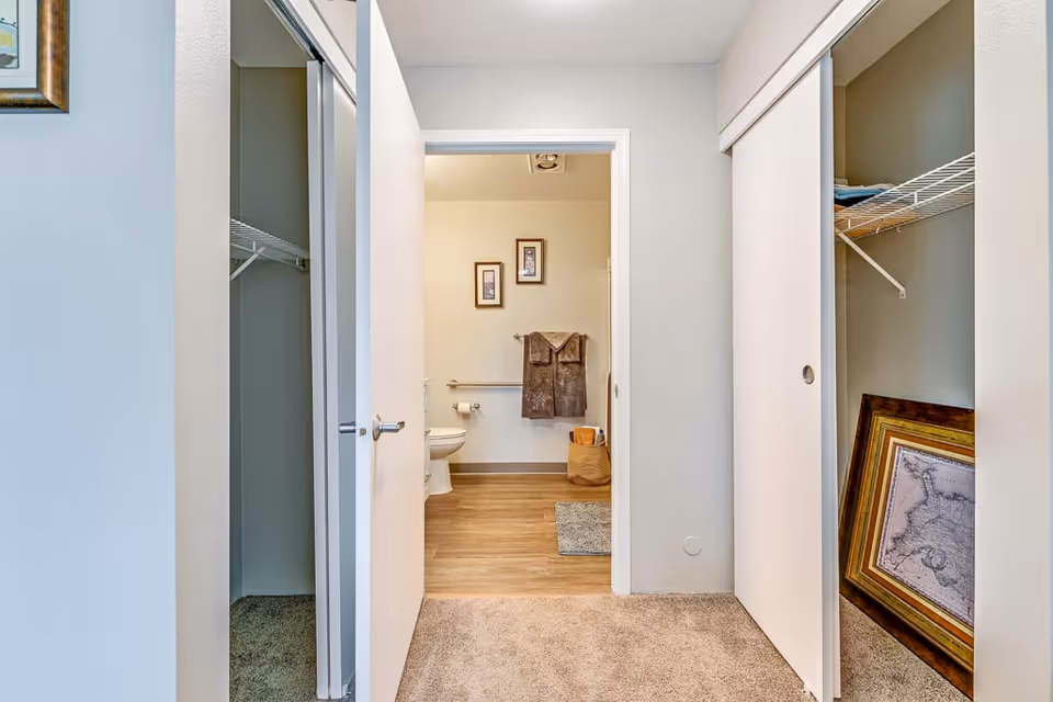 View through an open door into a bathroom with wooden flooring, a toilet, two framed pictures on the wall, a towel rack with two brown towels, and a woven basket on the floor. The foreground shows a carpeted area with two closets on either side, one with a sliding mirror door and the other with a sliding white door partially open revealing a framed picture inside.