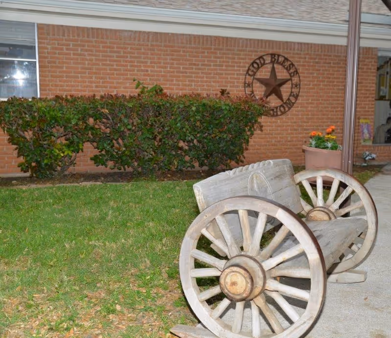 A rustic wooden bench made from wagon wheels sits on a grassy lawn next to a concrete walkway. Behind the bench is a neatly trimmed hedge and a brick wall with a decorative metal star and the words 'GOD BLESS OUR HOME'. A potted plant with orange flowers is placed near the bench.