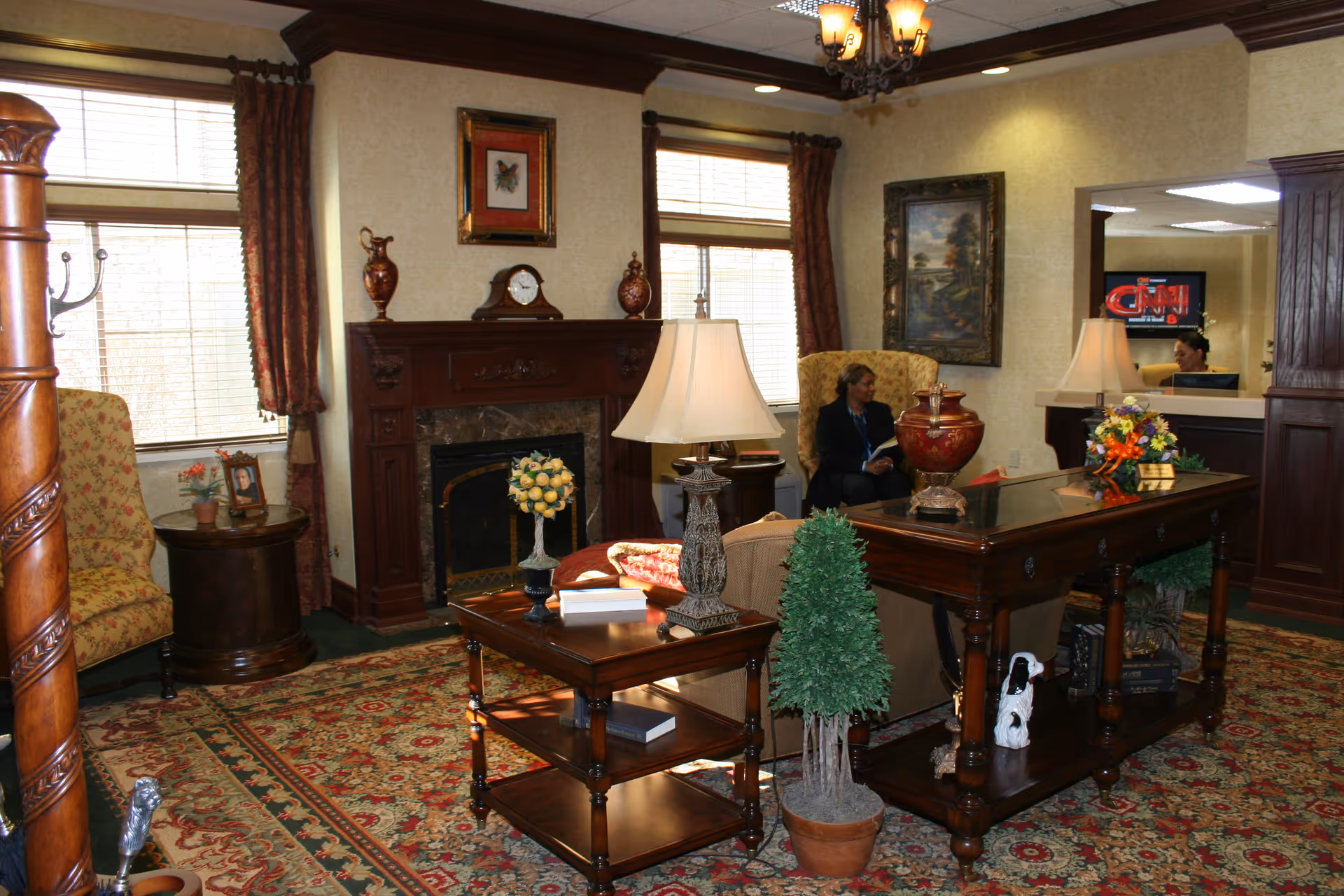 A cozy living room area in a nursing center featuring a wooden fireplace with decorative vases and a clock on the mantel. There are two large windows with curtains, a patterned rug covering the floor, and several pieces of wooden furniture including tables with lamps, a small potted plant, and a decorative vase. Two women are seated in the background near a reception desk with a television screen displaying the CNN logo.