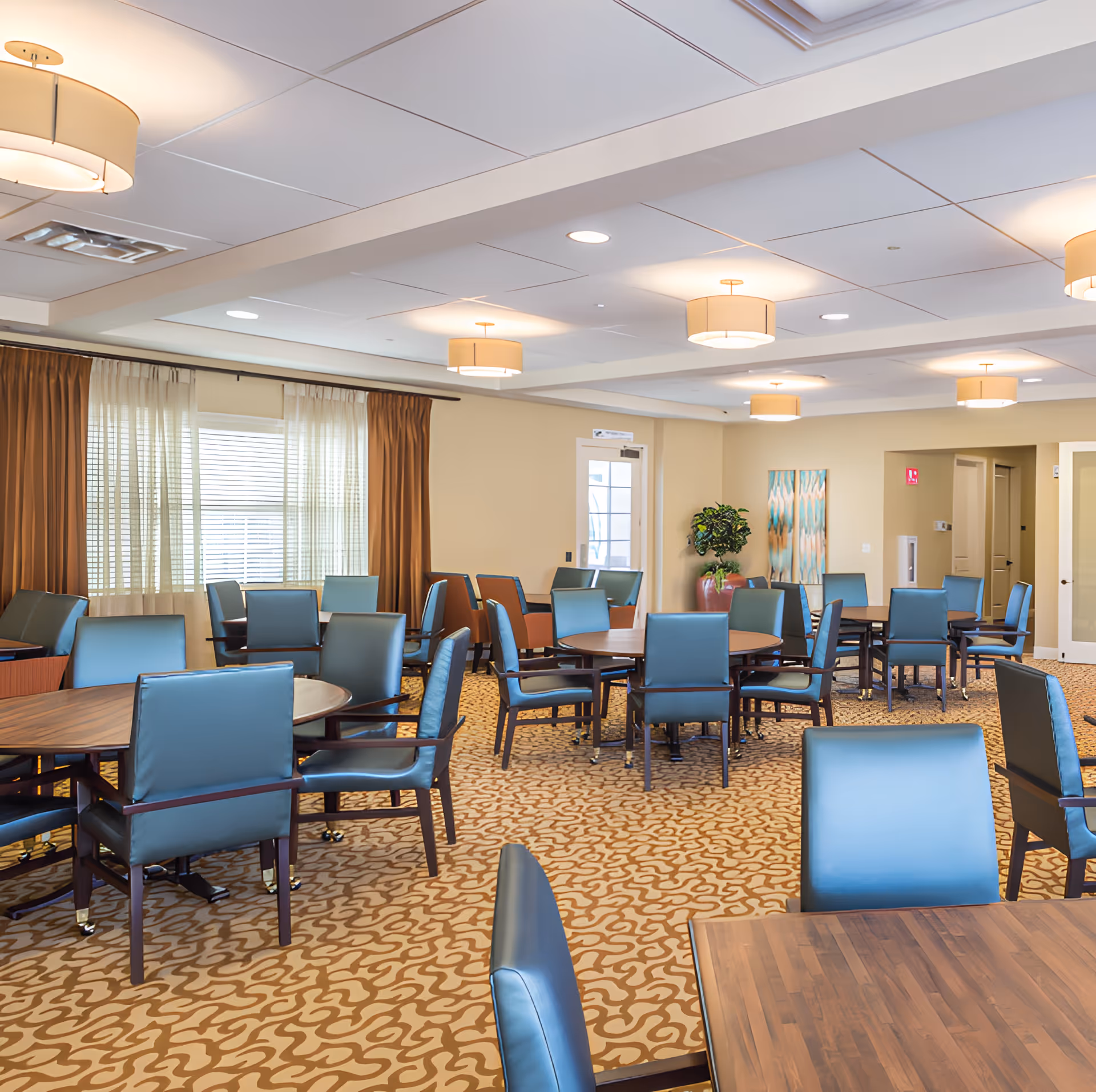 A spacious dining room with multiple round wooden tables surrounded by blue cushioned chairs. The room has beige walls, patterned carpet, large windows with sheer and brown curtains, and ceiling lights with round shades. There is a potted plant and abstract artwork on one wall.