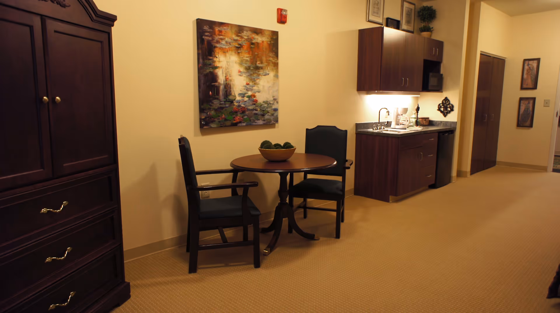 Interior view of a room with a small round wooden table and two dark wooden chairs with dark cushions. On the table is a decorative bowl with green moss balls. To the right is a kitchenette with dark wood cabinets, a sink, a coffee maker, and a mini refrigerator. On the left side is a large dark wooden cabinet with drawers. The walls are light beige with framed artwork and a fire alarm visible.