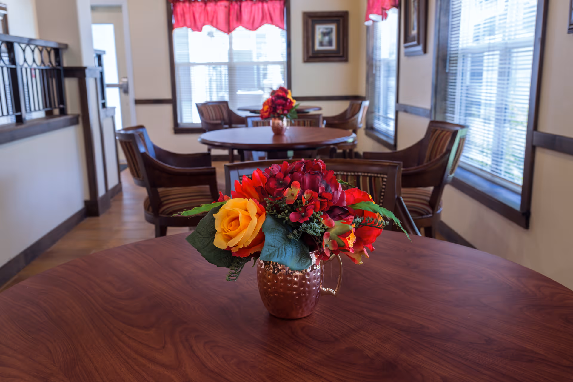 Interior view of a dining area with round wooden tables and cushioned chairs. Each table has a decorative flower arrangement in a copper vase. The room has large windows with blinds and red valances, allowing natural light to fill the space.