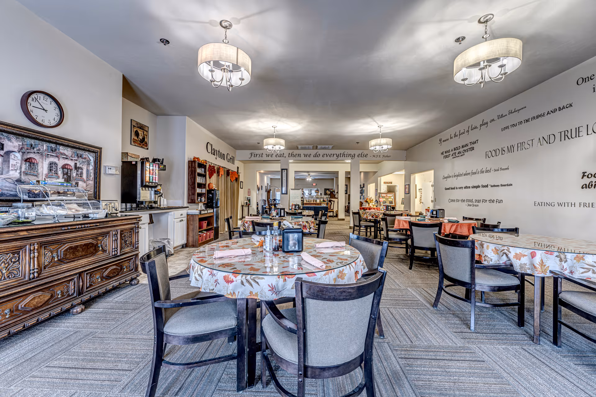 A bright dining room in Clayton Westlake Independent Living Senior Residences featuring round and square tables covered with floral tablecloths and set with napkins. The room has modern light fixtures hanging from the ceiling, a large clock on the wall, and a wooden sideboard with a display case. Inspirational food-related quotes are displayed on the walls, and there is a beverage station and buffet area labeled 'Clayton Grill' in the background.