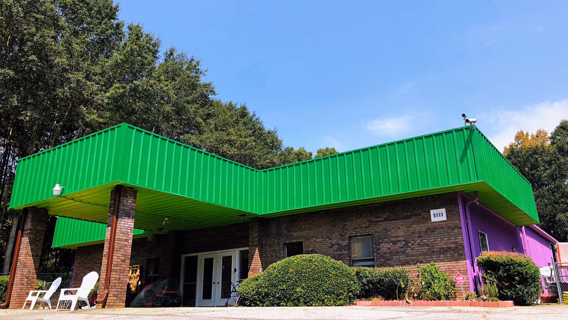 Front exterior of a low brick building with a bright green canopy and purple siding, surrounded by bushes and trees.