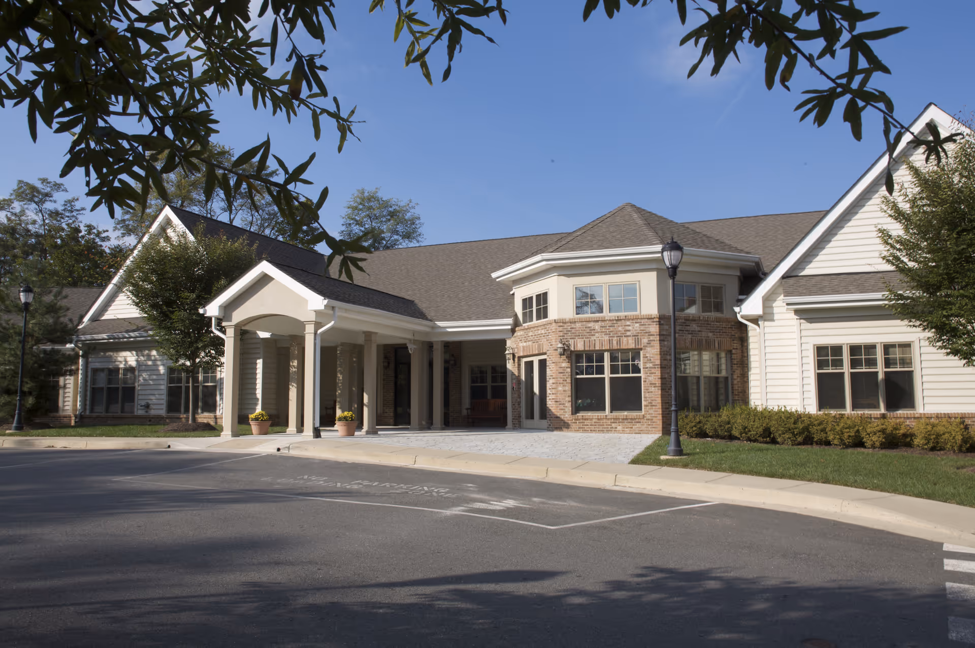 Entrance of a single-story brick and siding senior living building with a covered portico, lamp posts, and landscaped grounds under a clear blue sky.