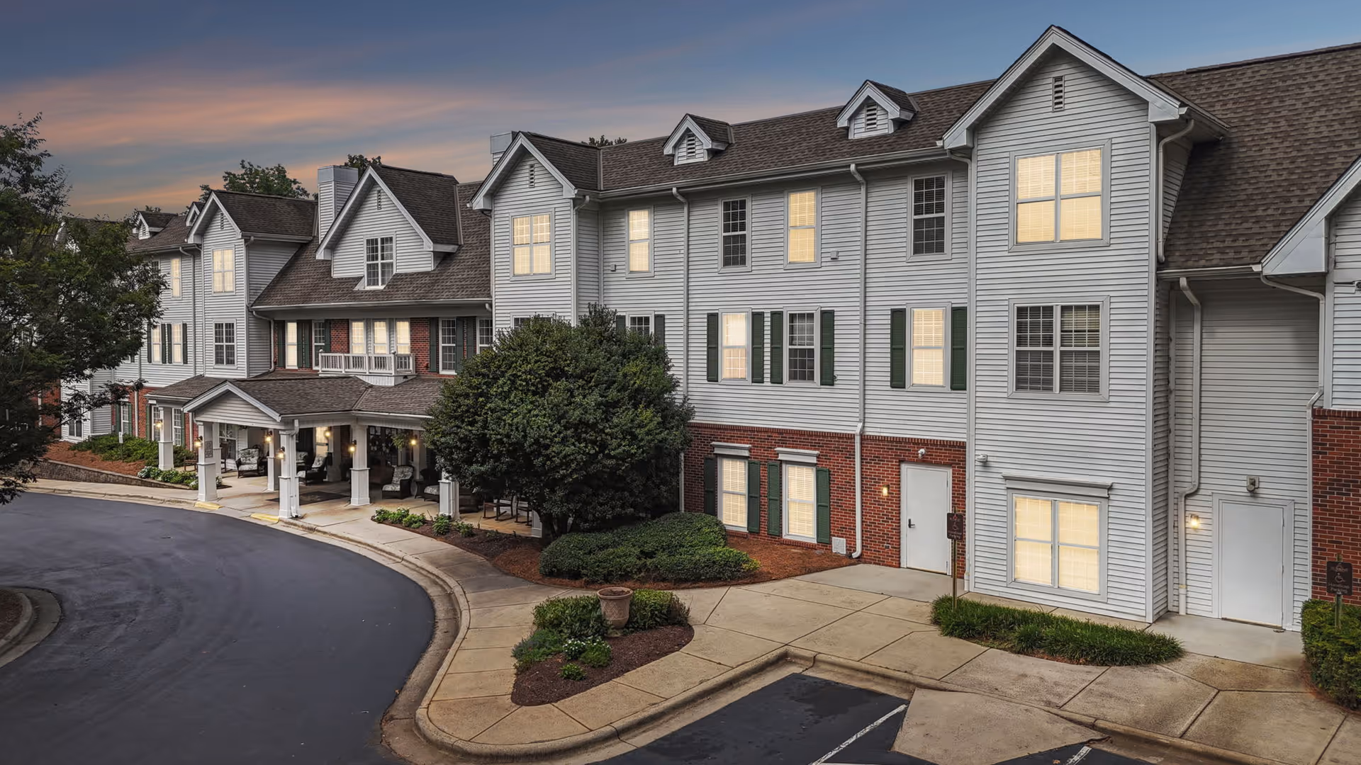 Exterior view of a multi-story senior living facility building at dusk with lit windows, a covered entrance with columns, landscaped bushes, and a curved driveway.