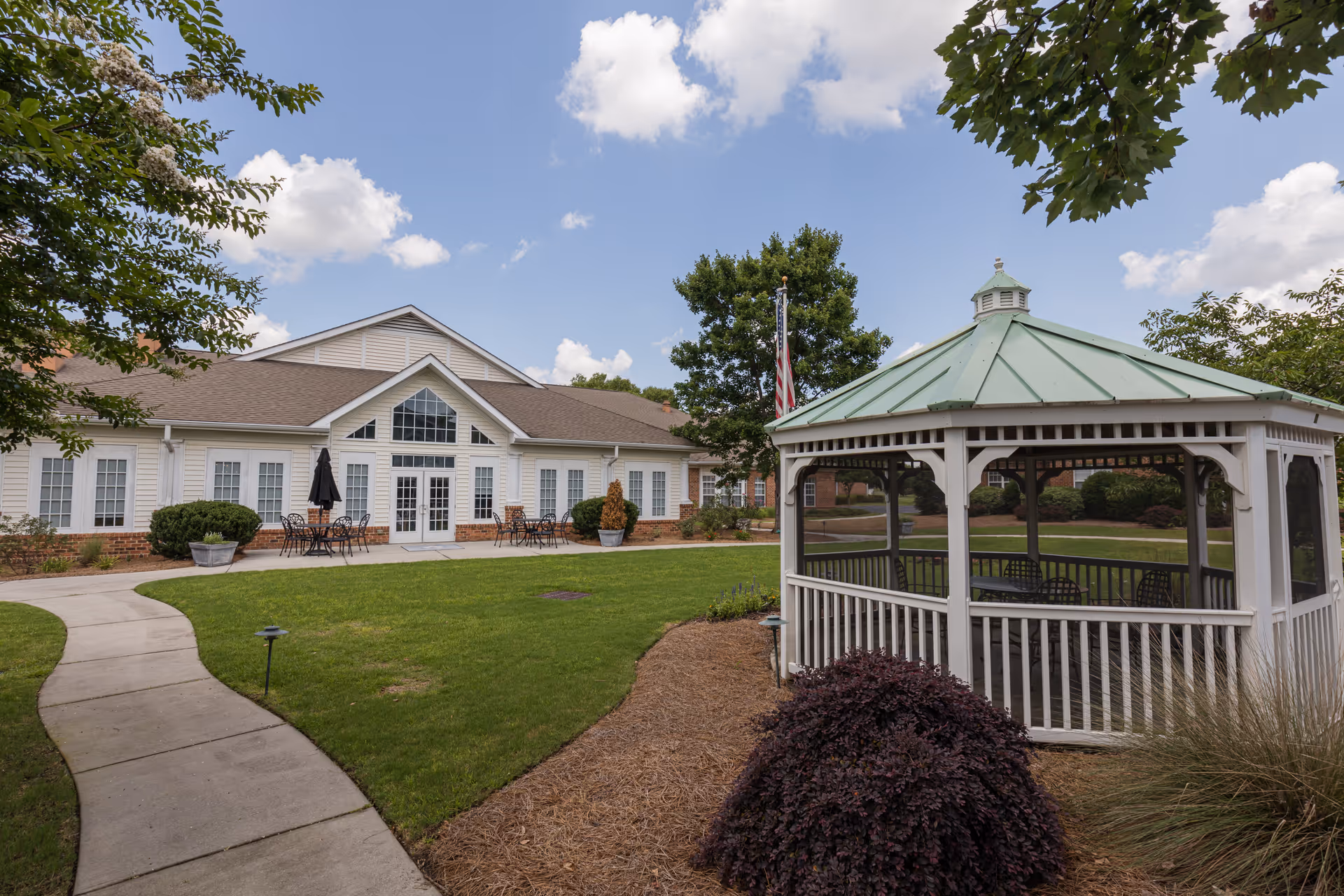 Outdoor view of Wildewood Downs facility showing a white gazebo with a green roof on the right, a curved concrete walkway, green lawn, and a building with multiple windows and patio tables with umbrellas under a partly cloudy sky.