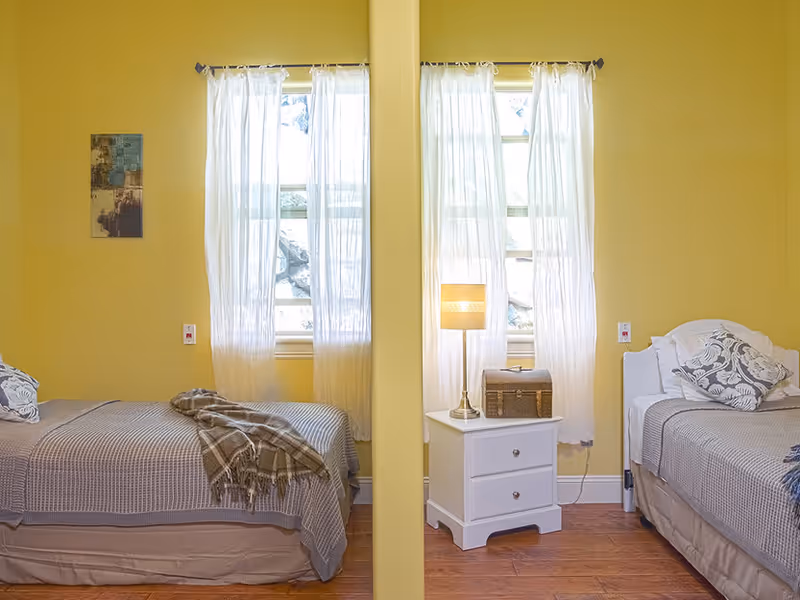Two twin beds in a yellow-painted shared bedroom separated by a partial wall, with a central nightstand, lamp, and windows with sheer curtains.