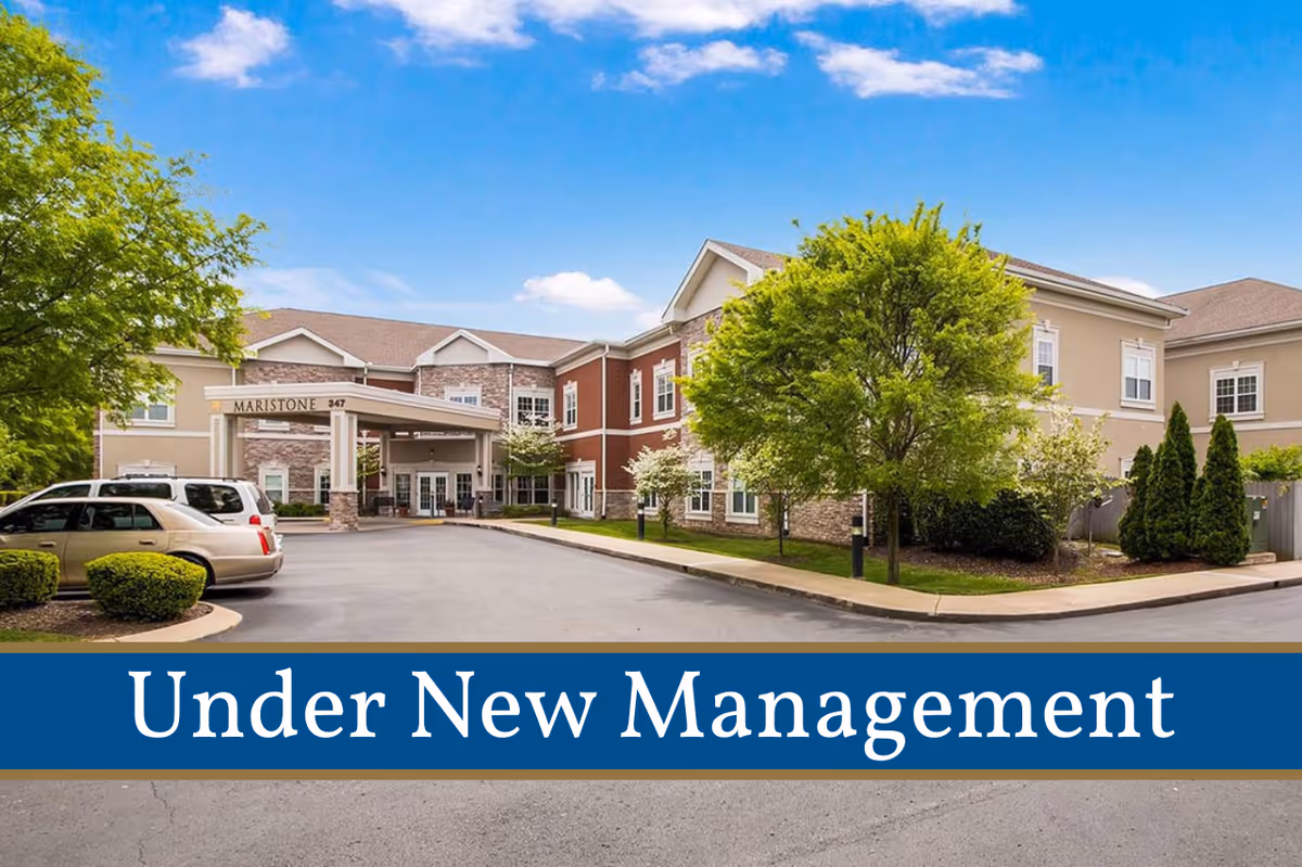 Exterior front entrance of the Maristone of Franklin senior living building with a driveway, parked cars, and landscaping, and an 'Under New Management' banner at the bottom.