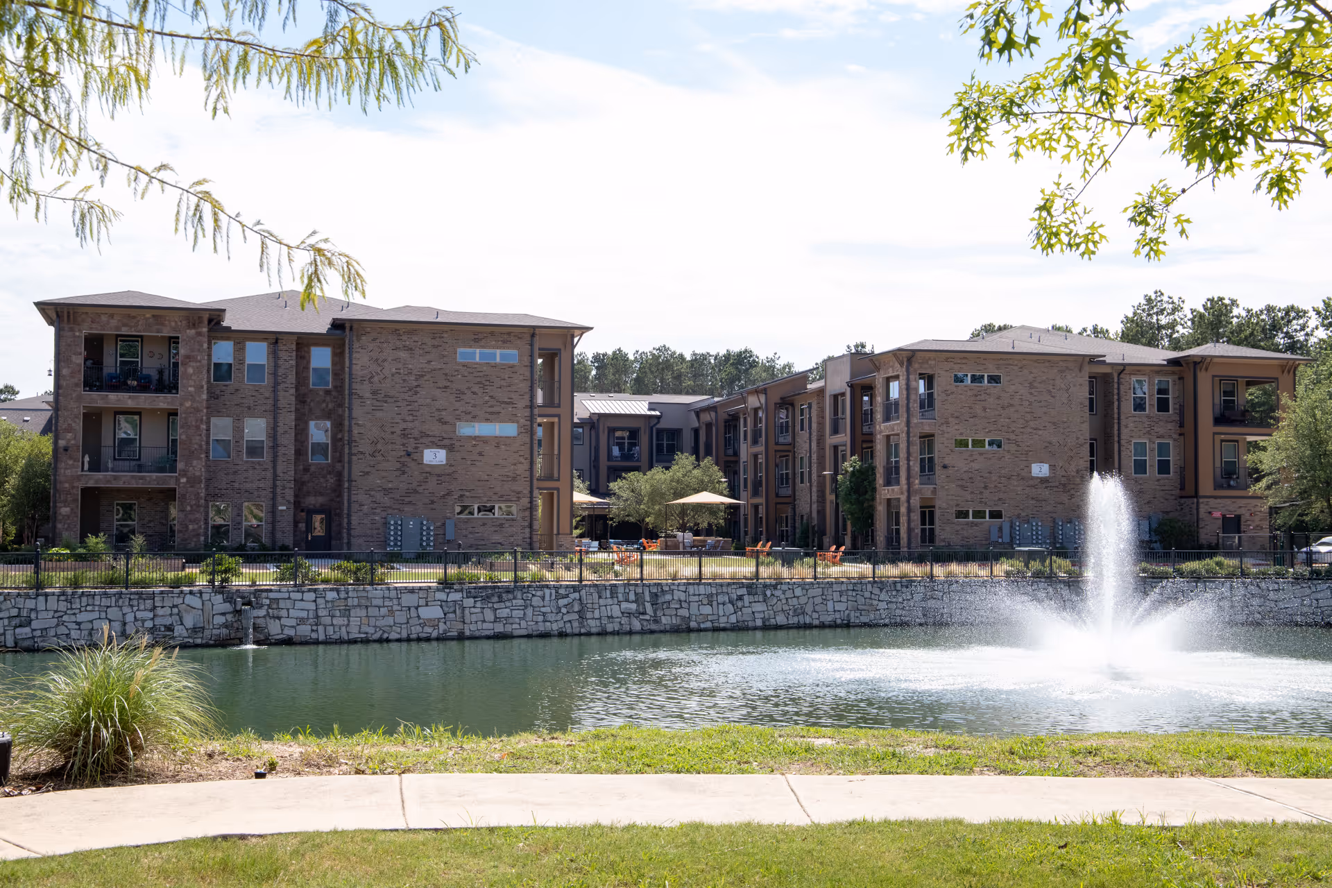 View of a senior living facility with two three-story brick buildings facing a pond with a water fountain in the center. There is a stone retaining wall along the pond and a grassy area with a sidewalk in the foreground. Trees with green leaves frame the top of the image.