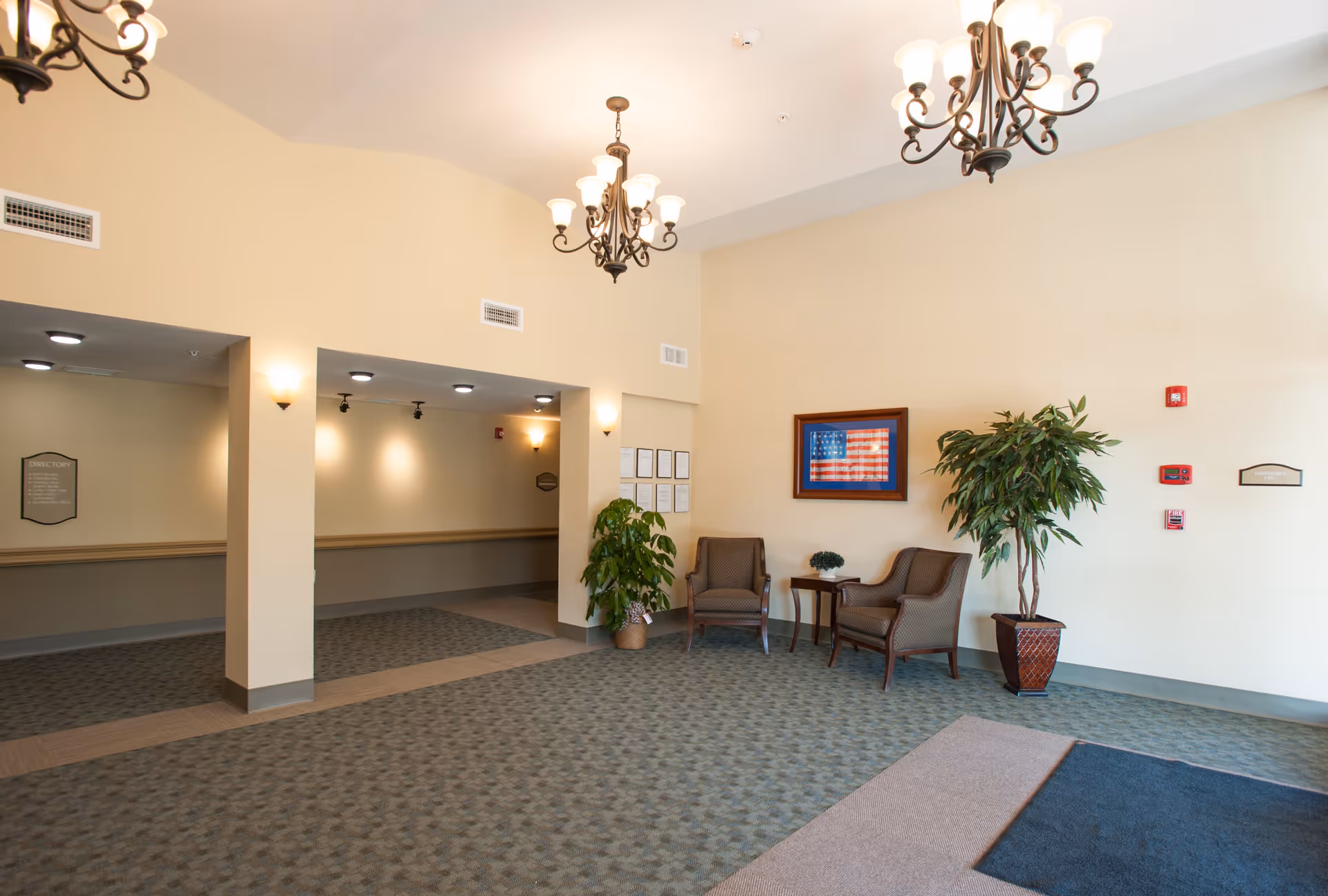 A spacious senior living facility lobby area with beige walls and carpeted floors. The room features two chandeliers hanging from the ceiling, two armchairs with a small table between them, a large potted plant, and a framed American flag on the wall. There are wall-mounted lights and a directory sign visible in the hallway area.