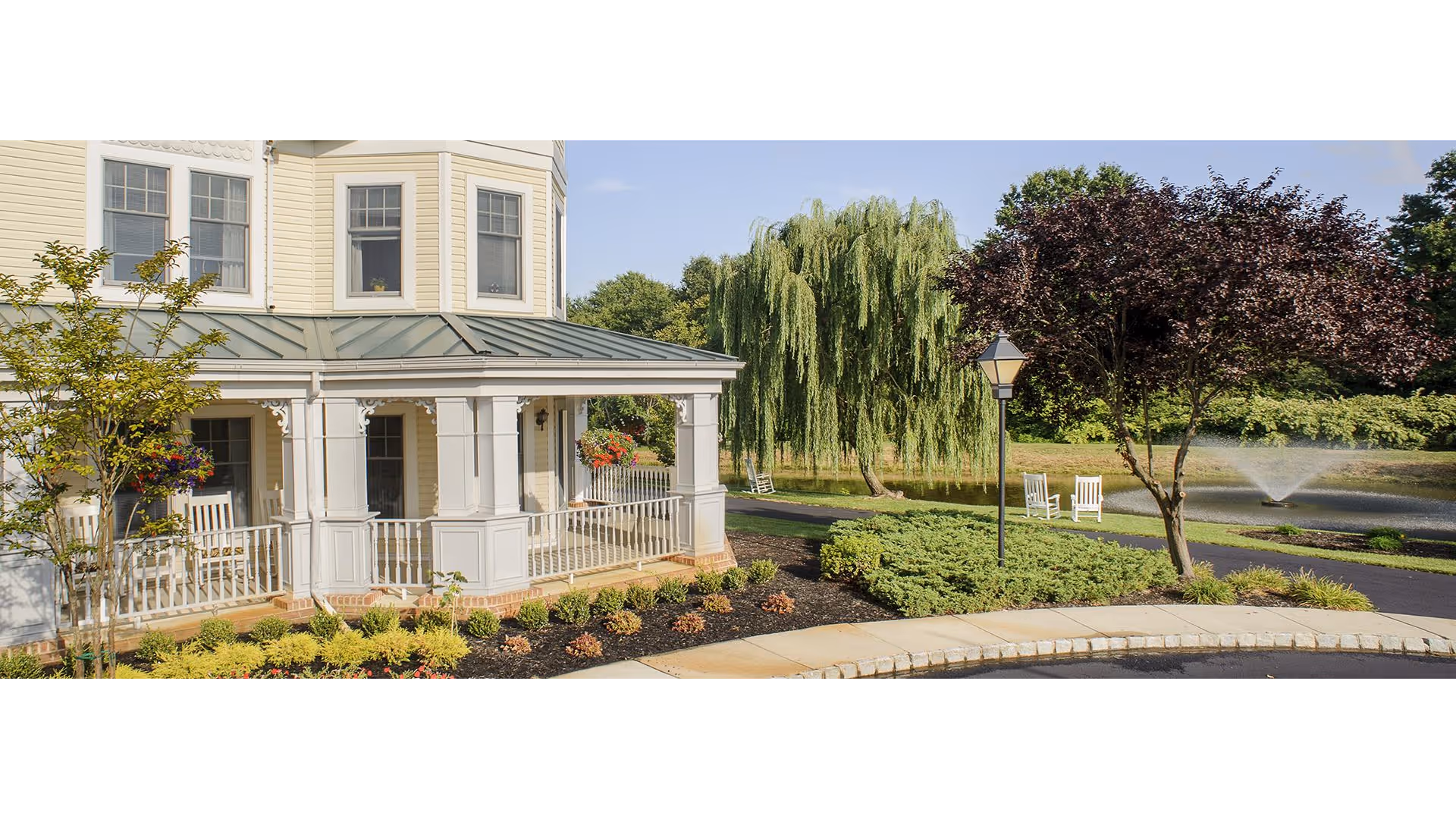 Exterior view of a senior living facility with a covered porch featuring white railings and columns. The building is light yellow with multiple windows. In the background, there is a pond with a fountain, a weeping willow tree, other trees, and white rocking chairs along a paved pathway.